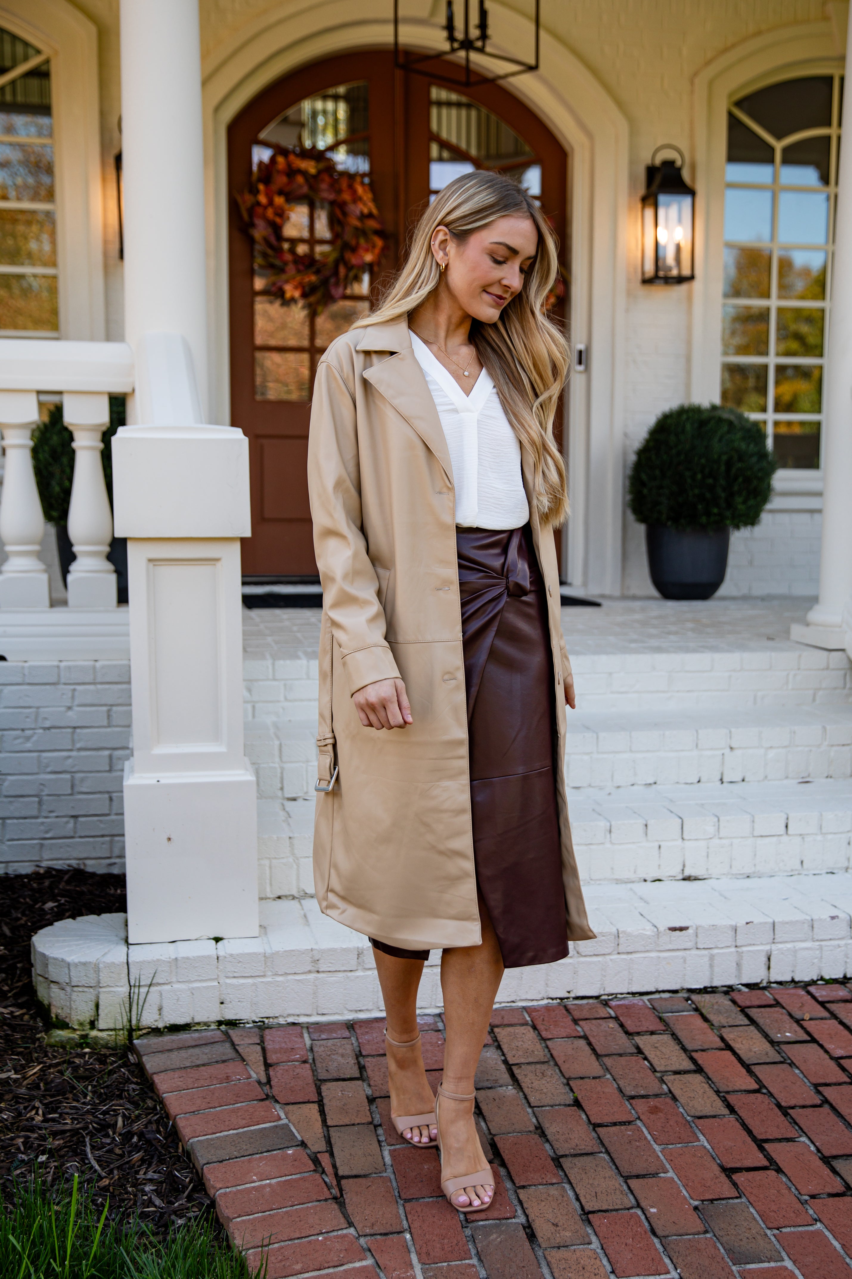 Woman in a beige trench coat and brown leather skirt standing on a porch.