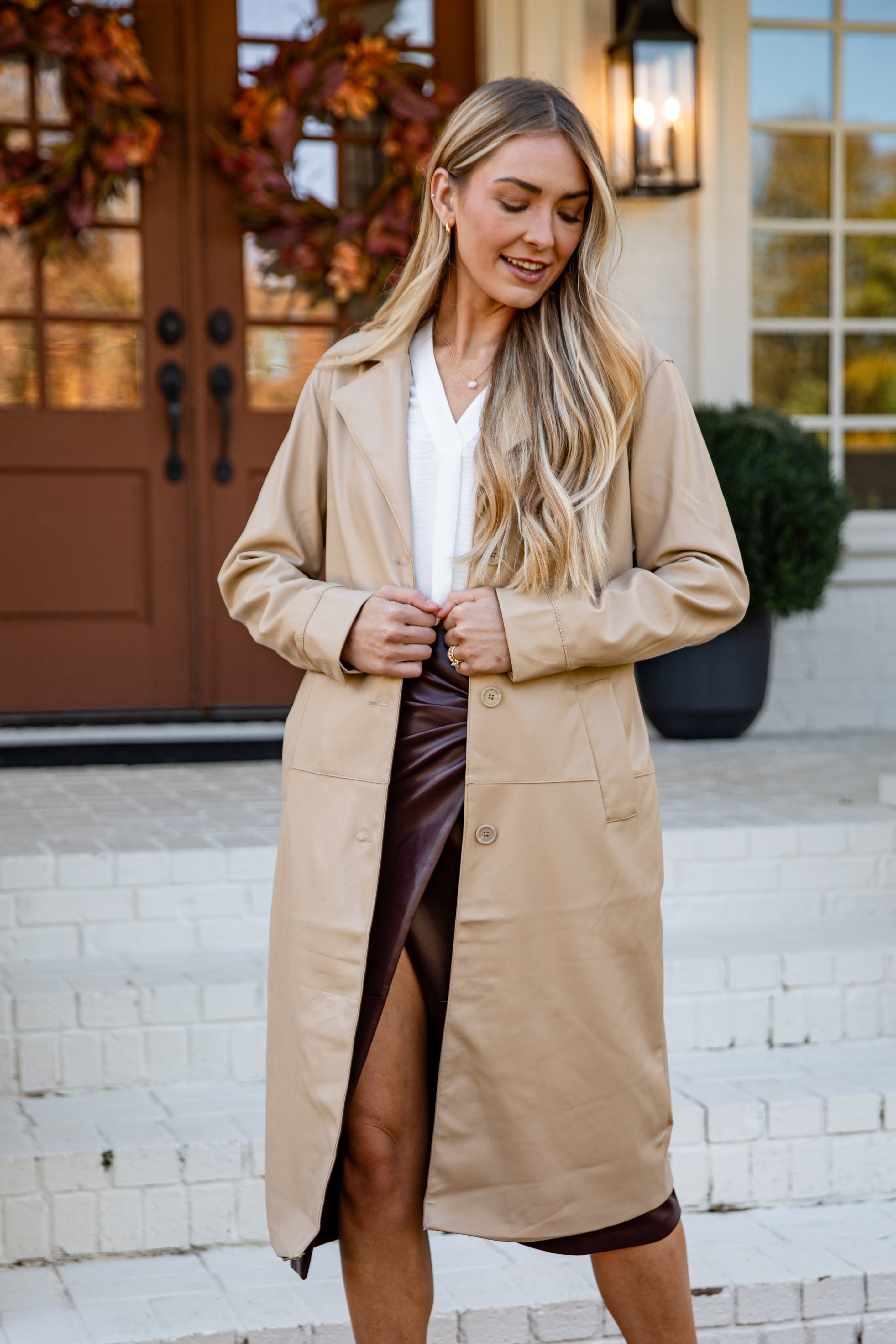 Woman wearing a beige trench coat standing outside a house.