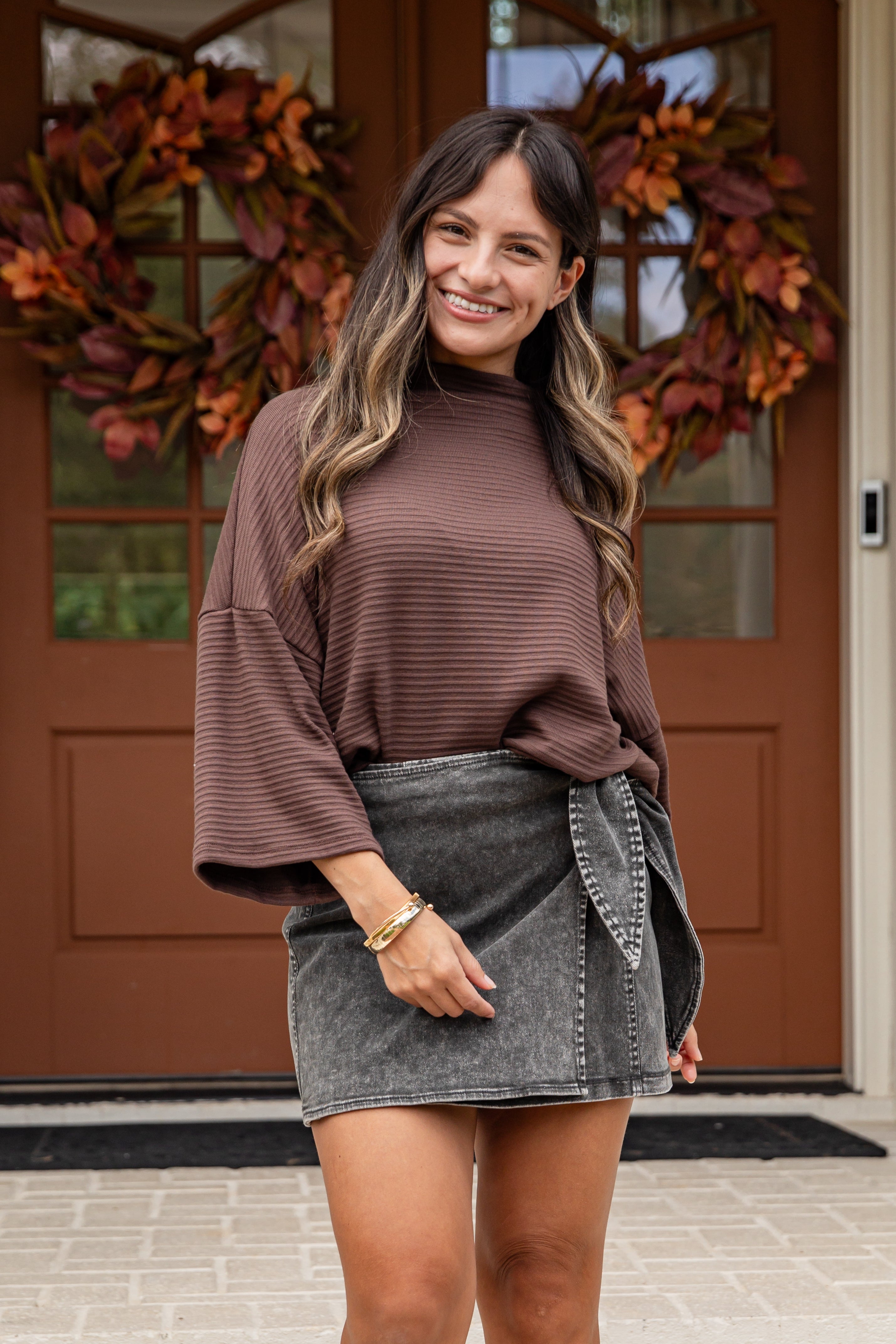 Woman wearing a brown ribbed sweater and denim skirt standing in front of a door with autumn wreaths.