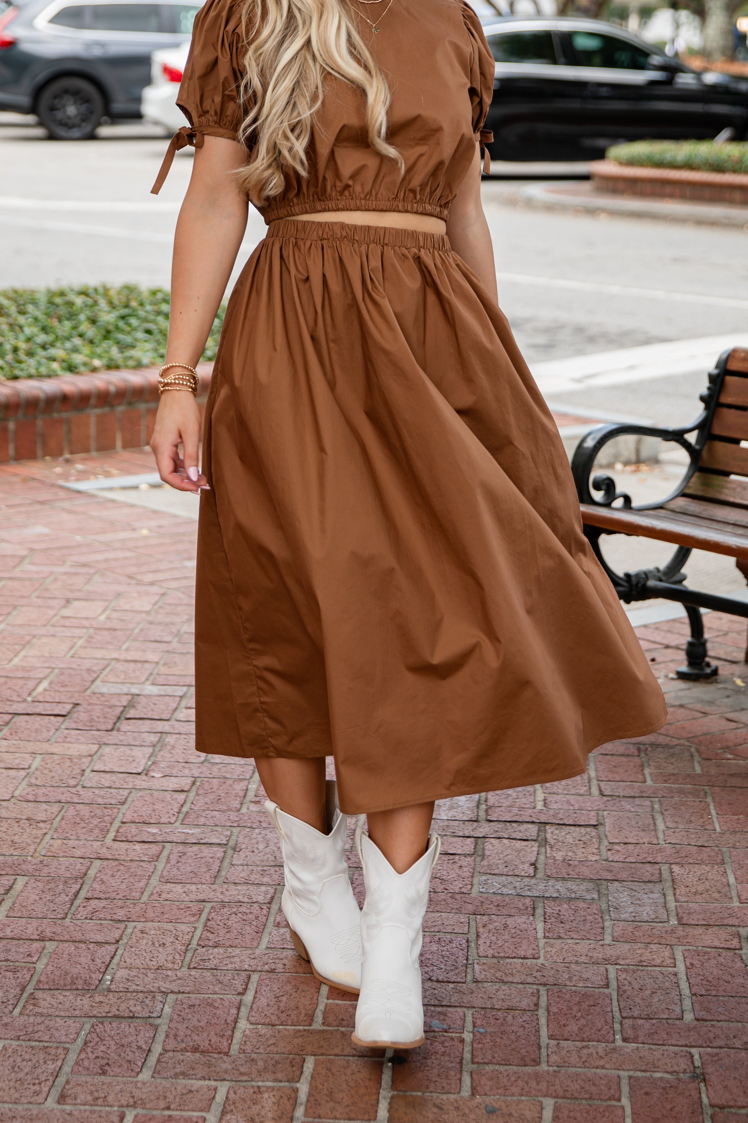 Woman in a brown dress and white boots walking on a sidewalk.