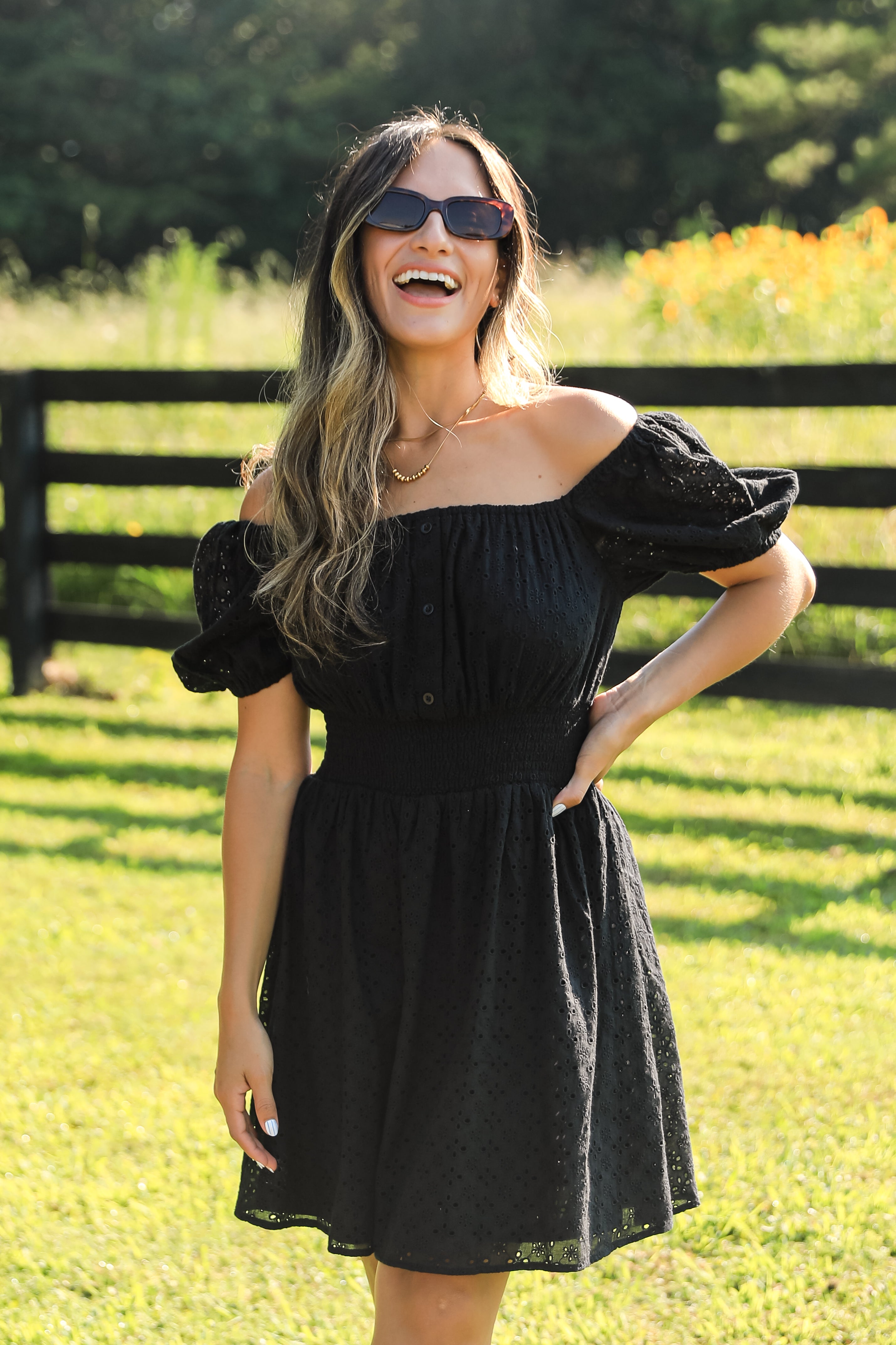 Woman in a black dress standing in a grassy field with a wooden fence and flowers in the background.