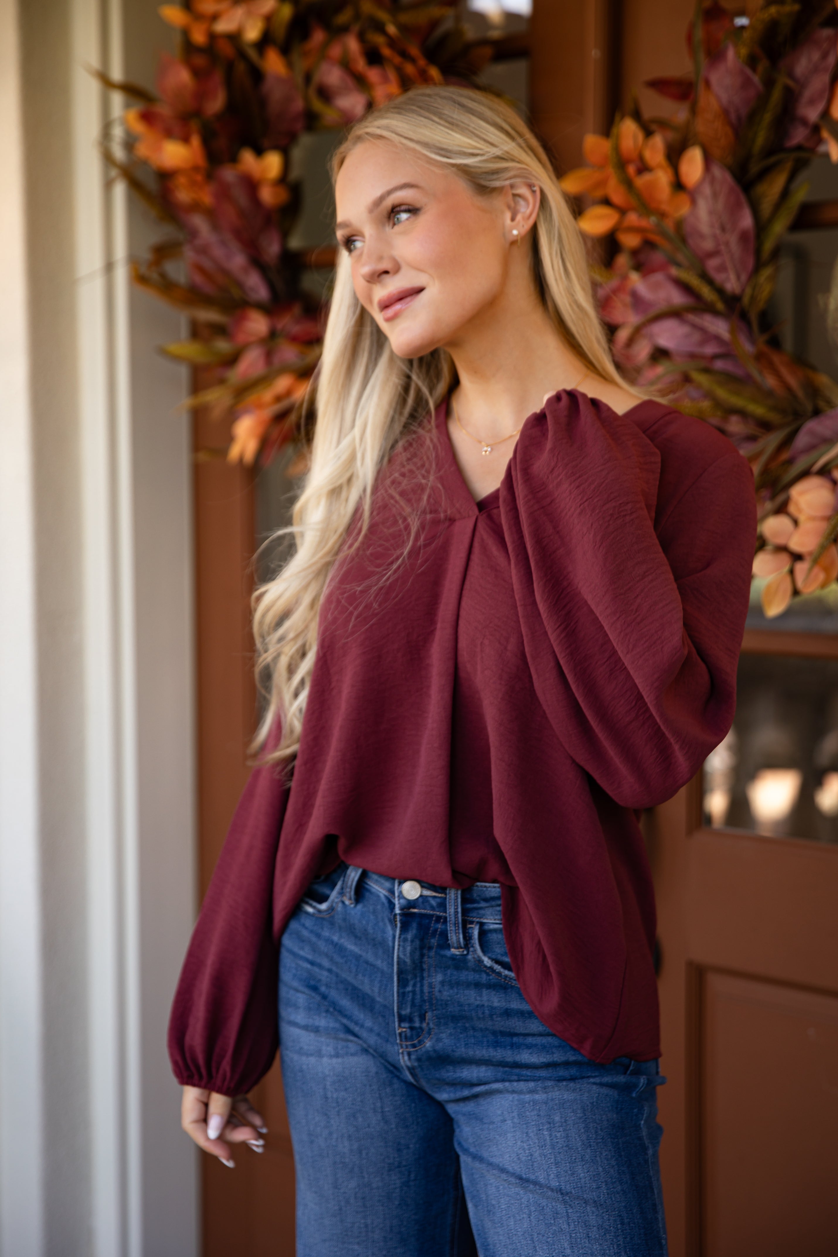 Woman wearing a burgundy blouse and blue jeans standing in front of autumn-themed decor.