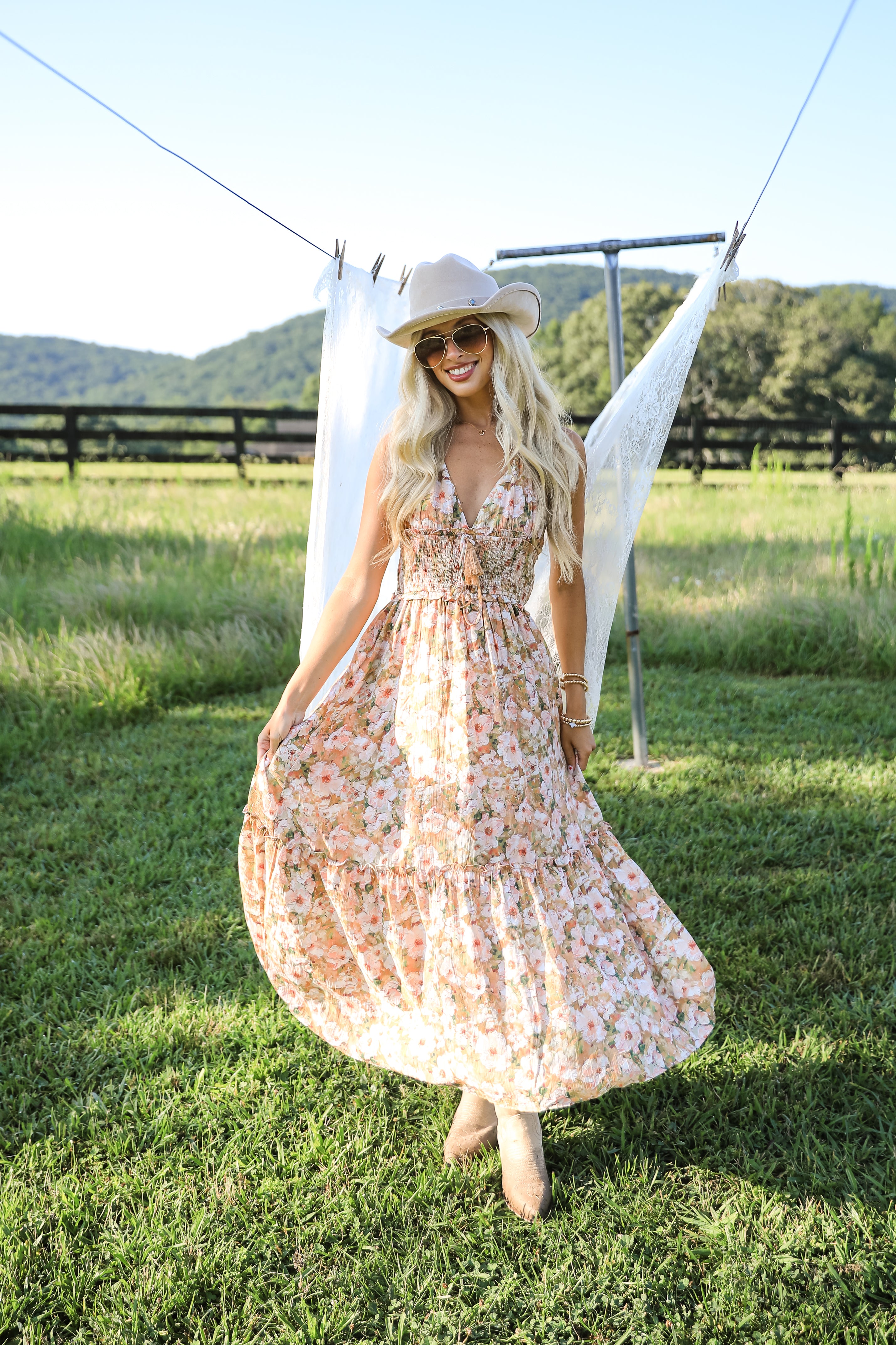 Woman in a floral dress and hat standing in a grassy field with mountains in the background