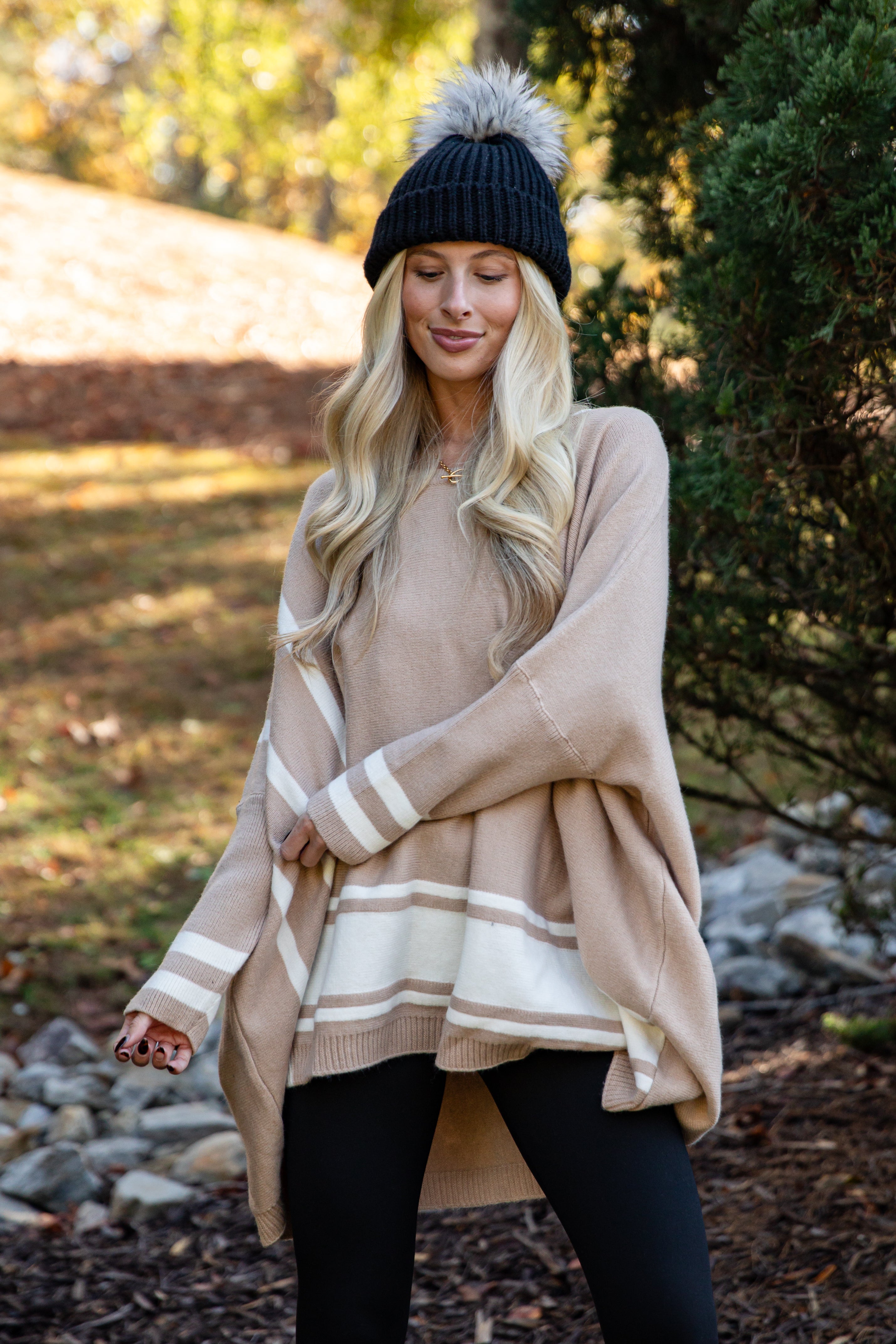 Woman wearing a beige poncho with white stripes outdoors near trees and rocks.