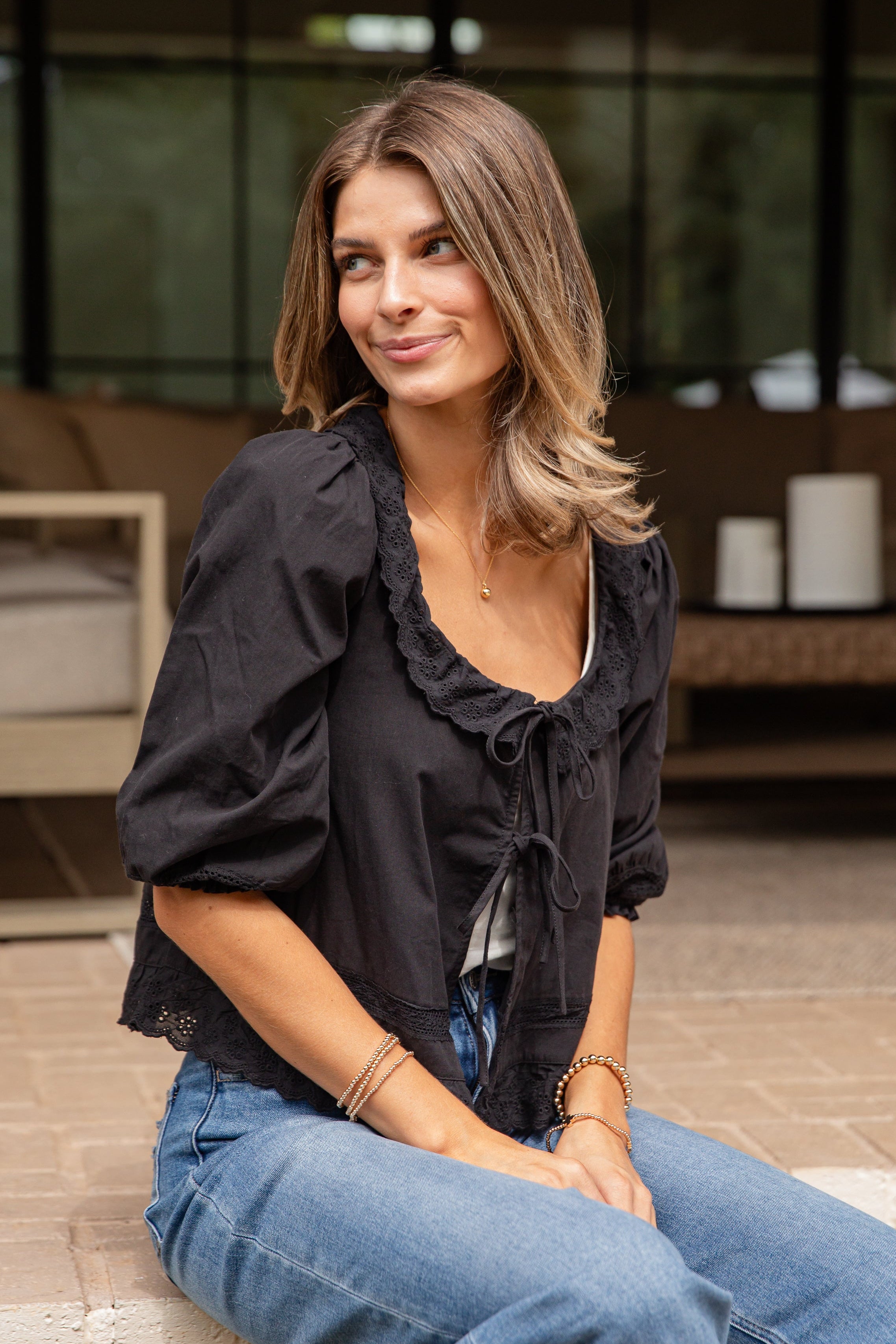 Woman sitting on a stone ledge wearing a black top and blue jeans, with a blurred indoor background.