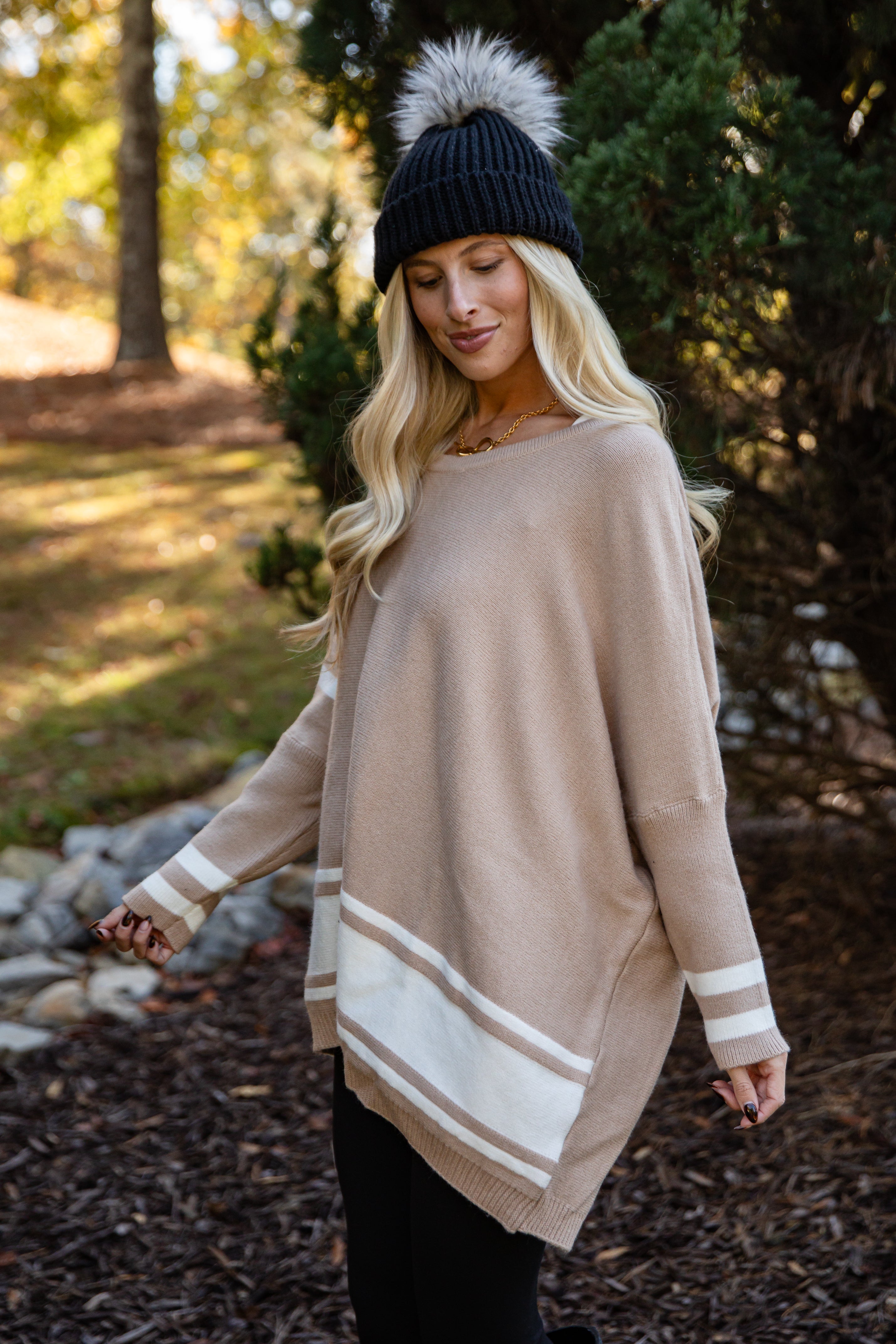 Woman wearing a beige sweater with white stripes and a black beanie in a forest setting