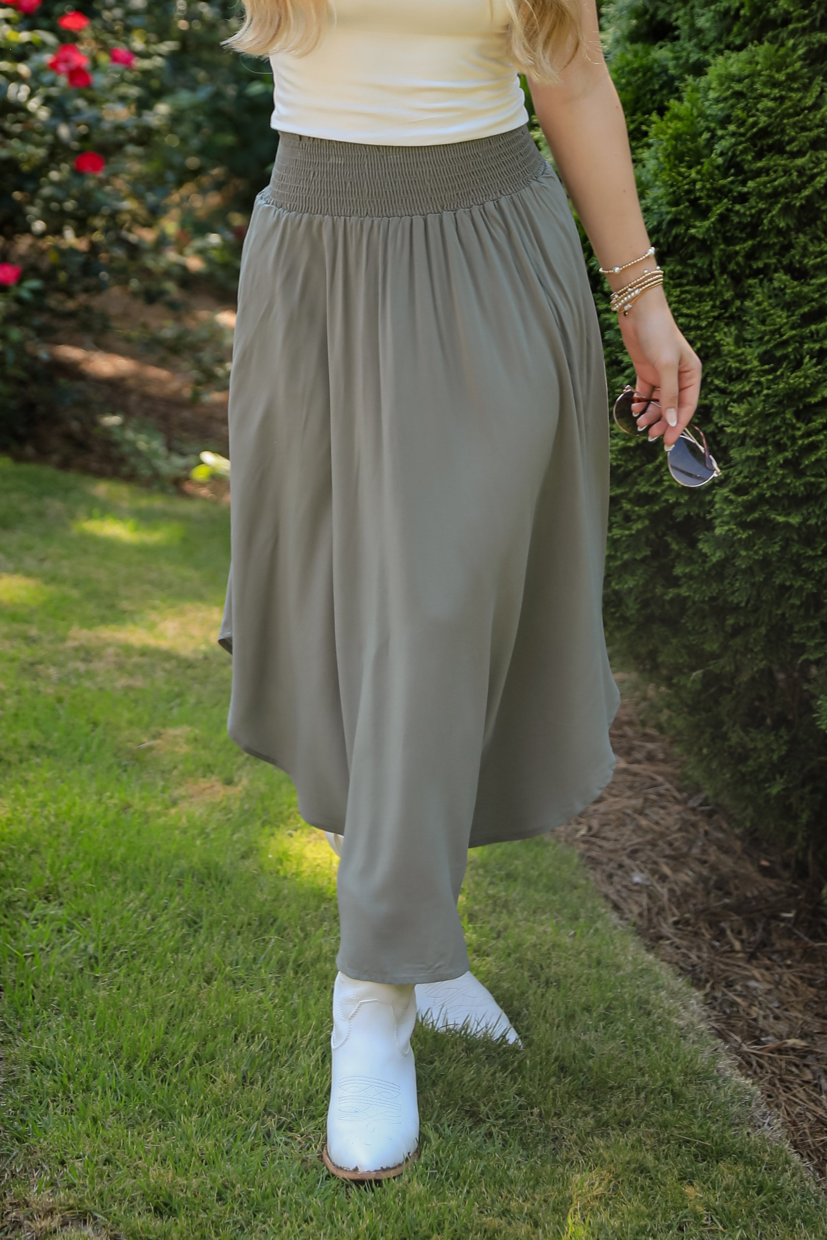 Person wearing a white top and gray skirt standing outdoors with greenery in the background