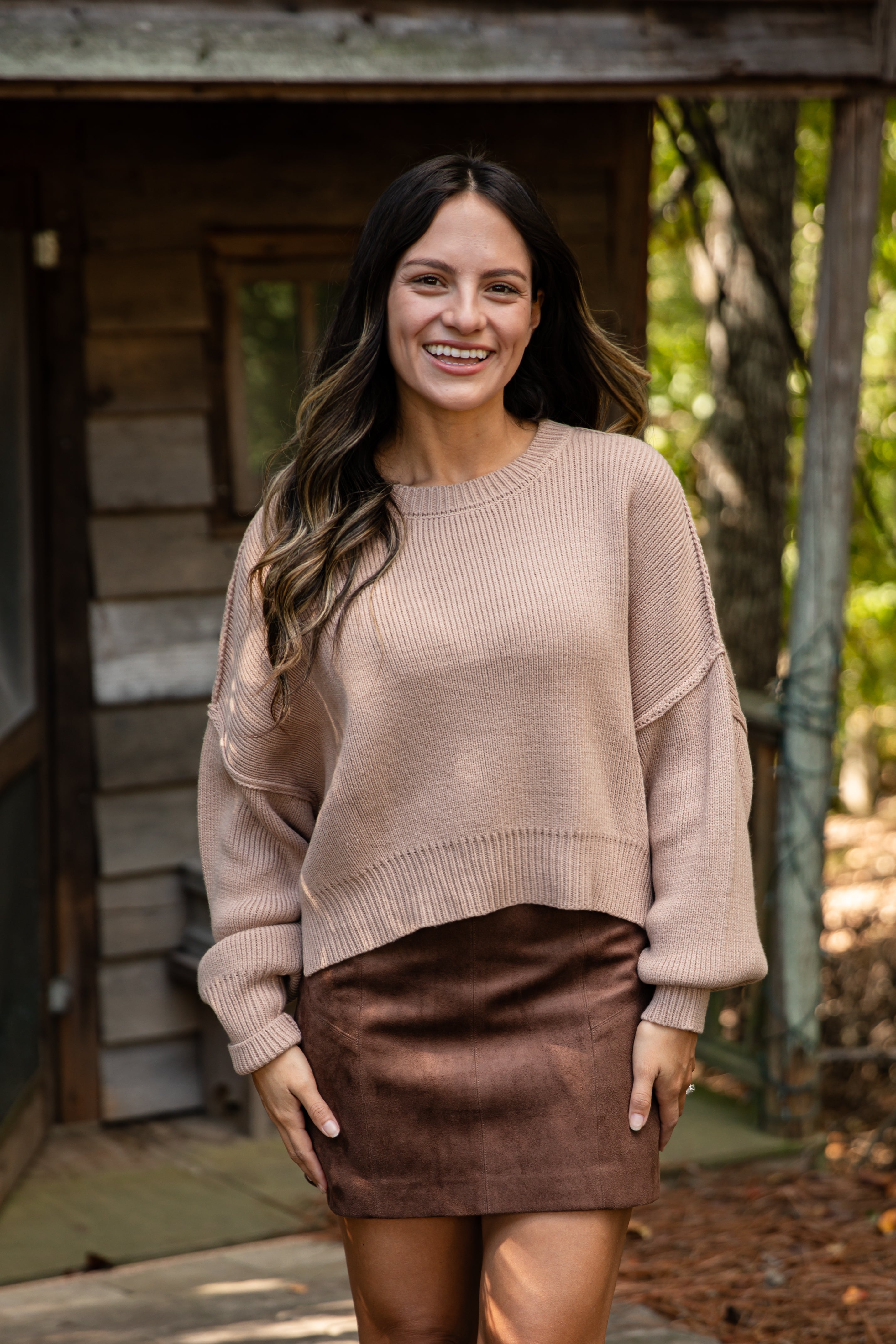 Woman wearing a beige sweater and brown skirt standing in front of a wooden cabin.