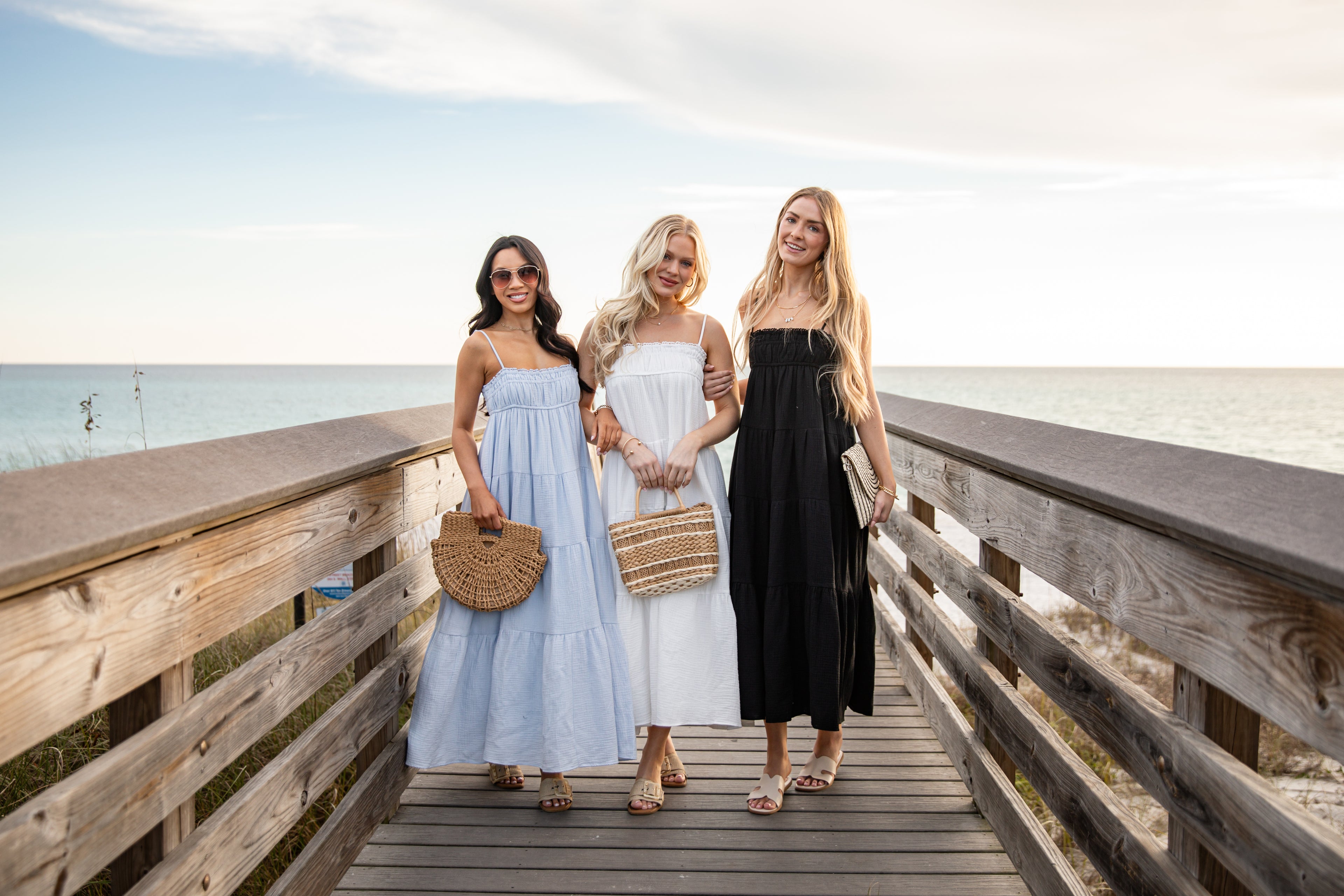 Three women standing on a wooden boardwalk by the ocean, wearing dresses.