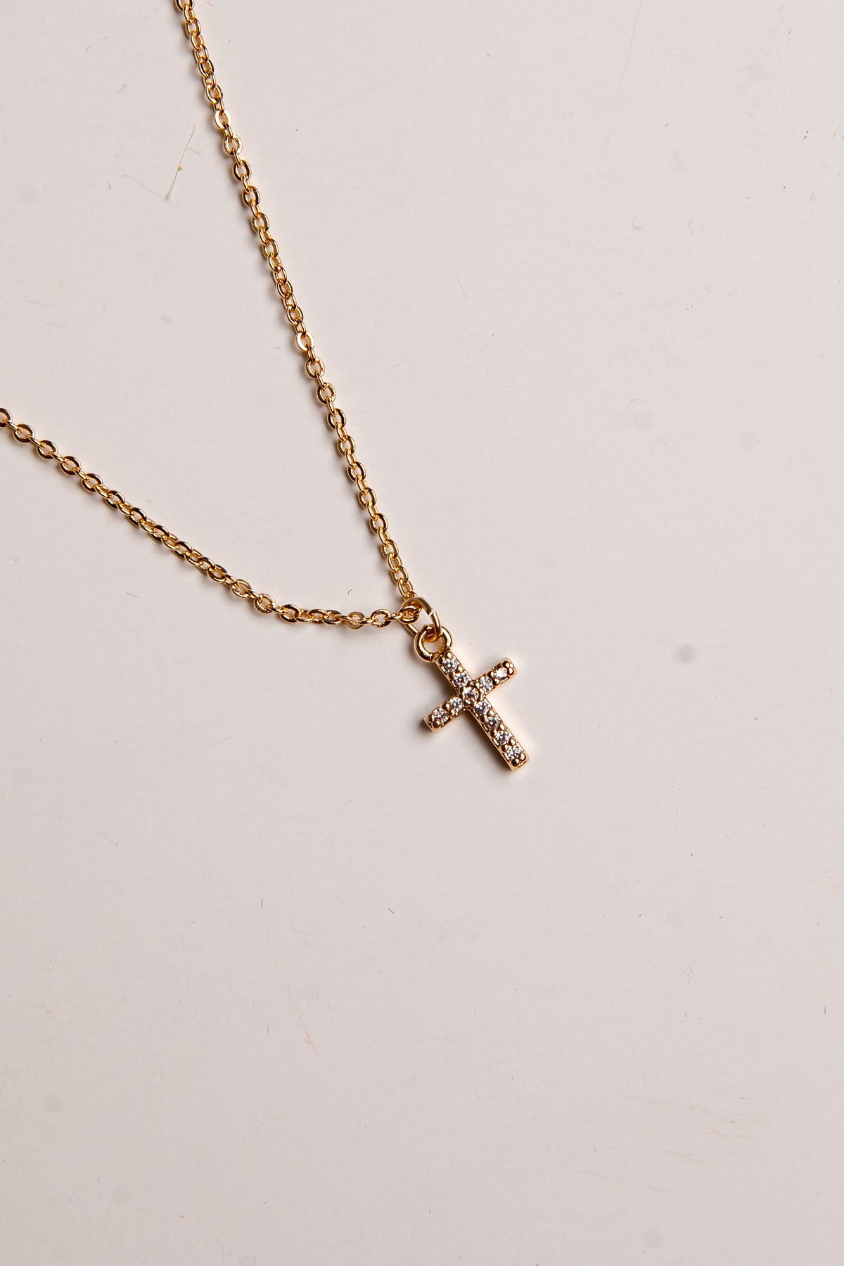 A delicate gold chain necklace featuring a small cross pendant adorned with tiny clear rhinestones, displayed on a light off-white background.