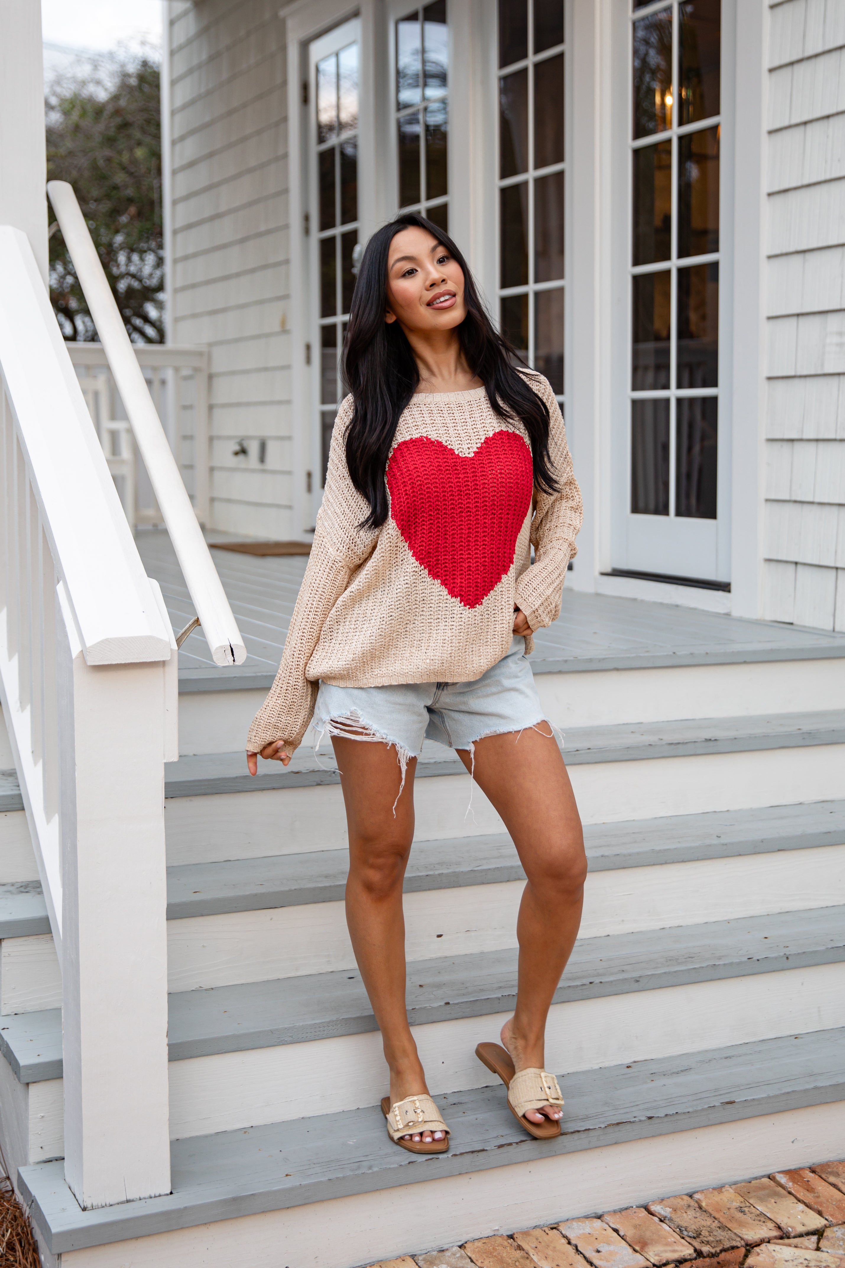 Woman wearing a beige sweater with a red heart design on steps outside a house.
