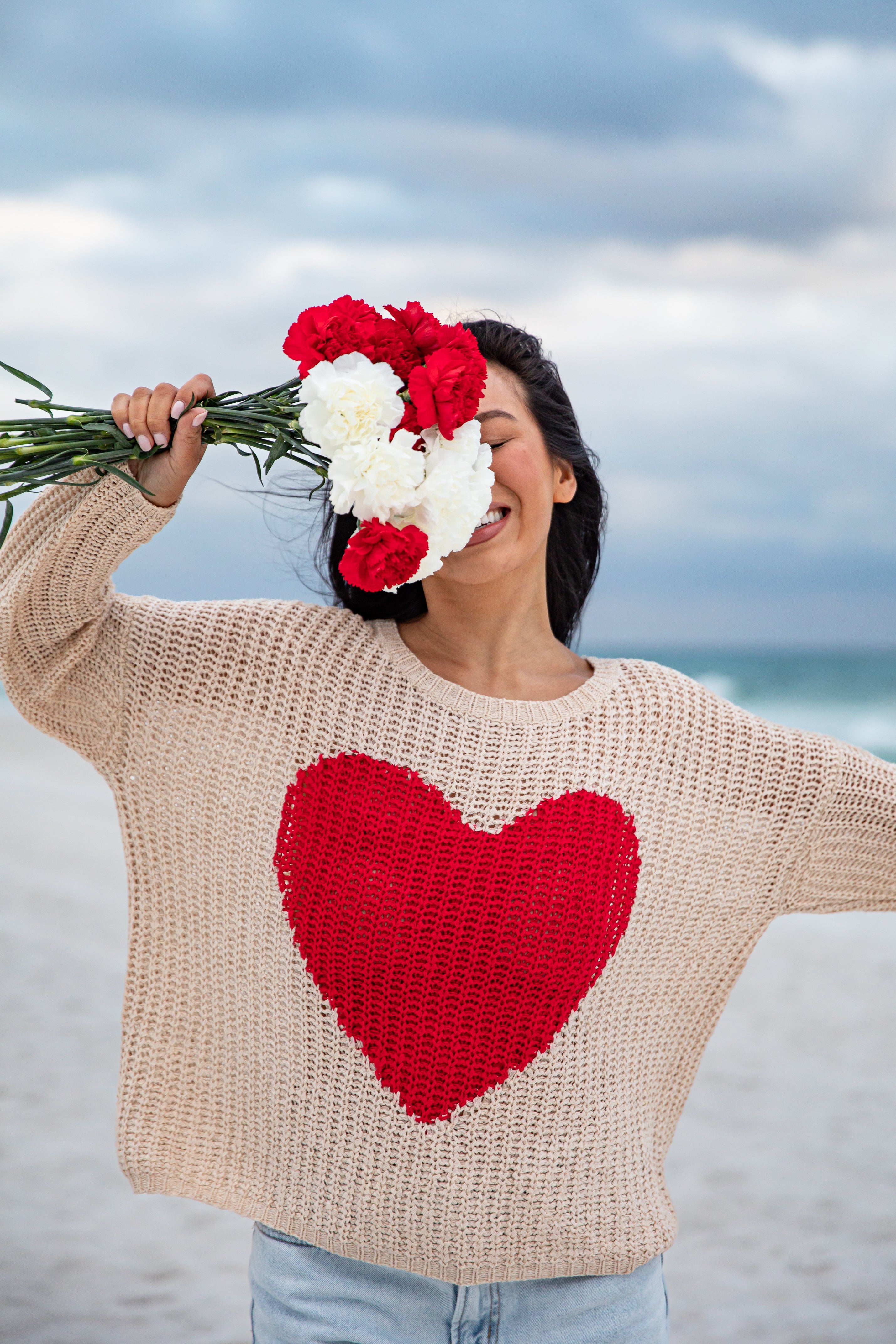Woman on a beach wearing a sweater with a red heart design, holding flowers.