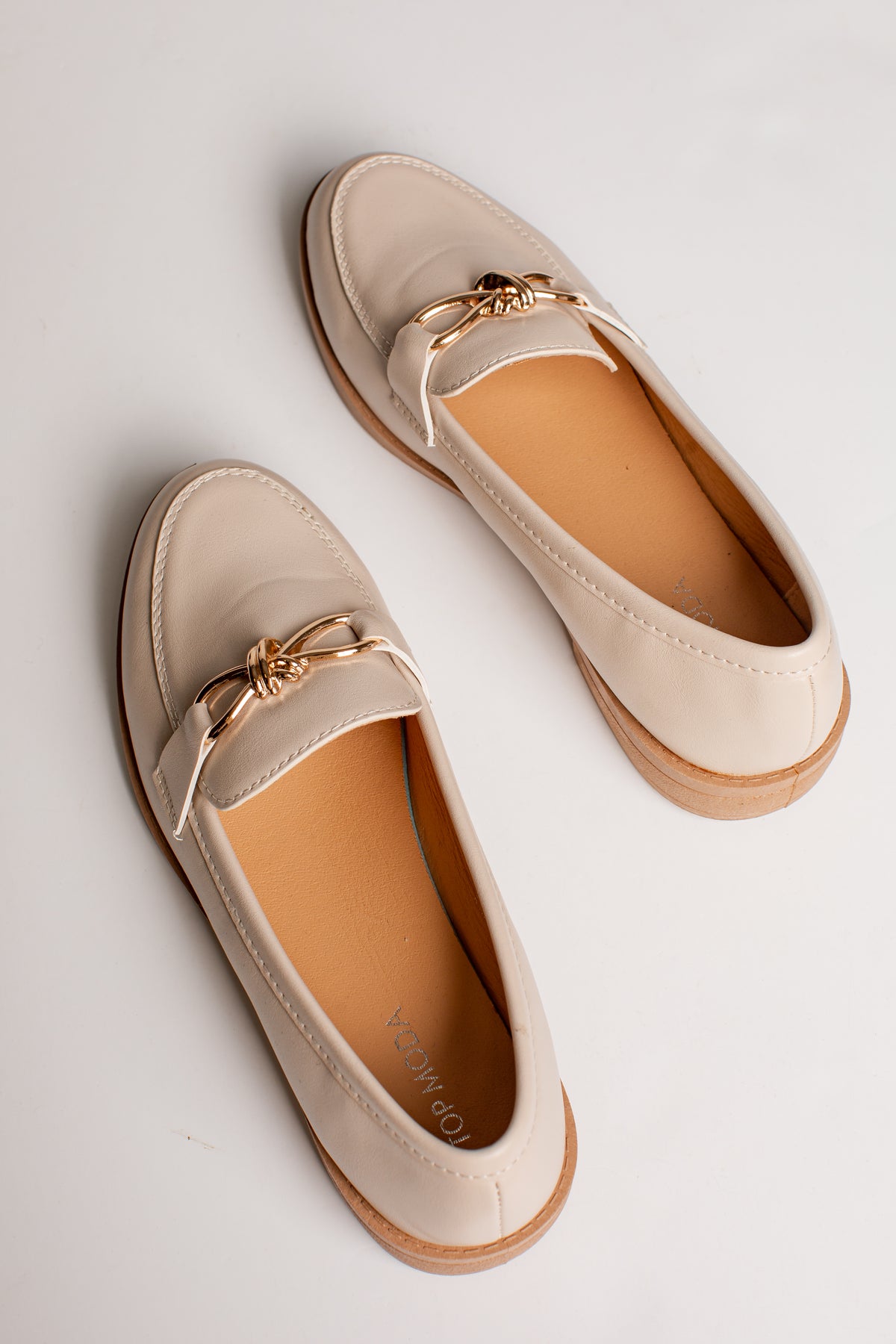 A pair of beige leather penny loafers with a gold-tone metal bow detail across the top, displayed on a clean white background.
