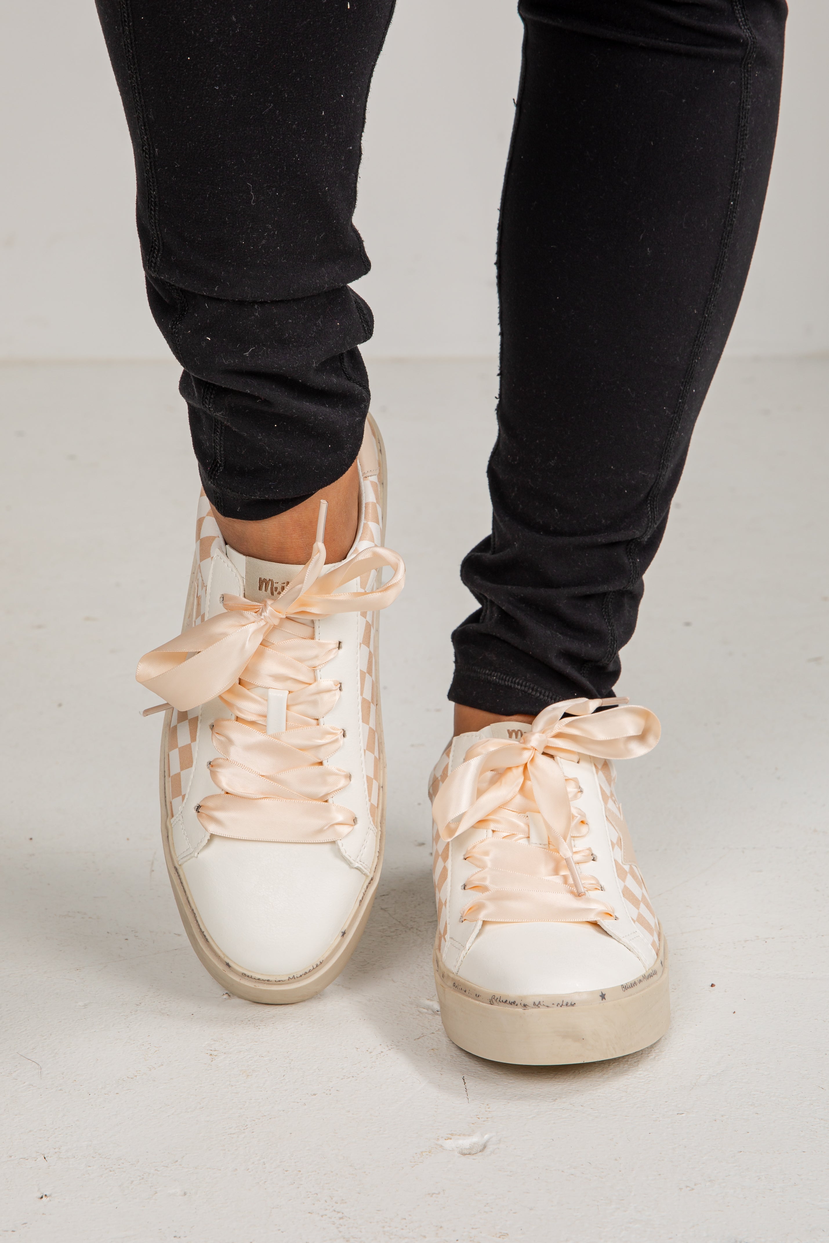White sneakers with pink laces worn with black pants on a light gray background