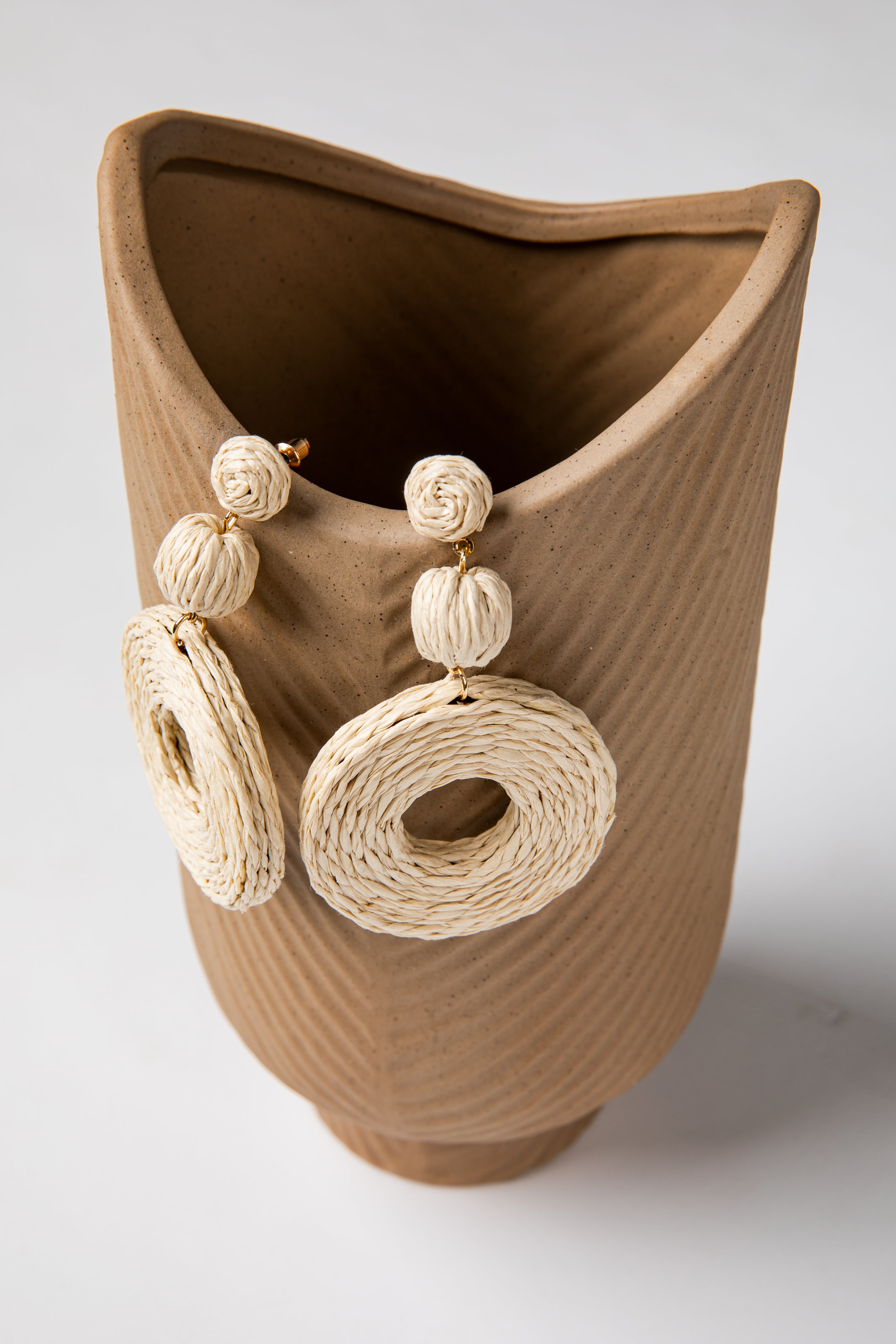 Brown ceramic vase with woven beige earrings attached on a white background
