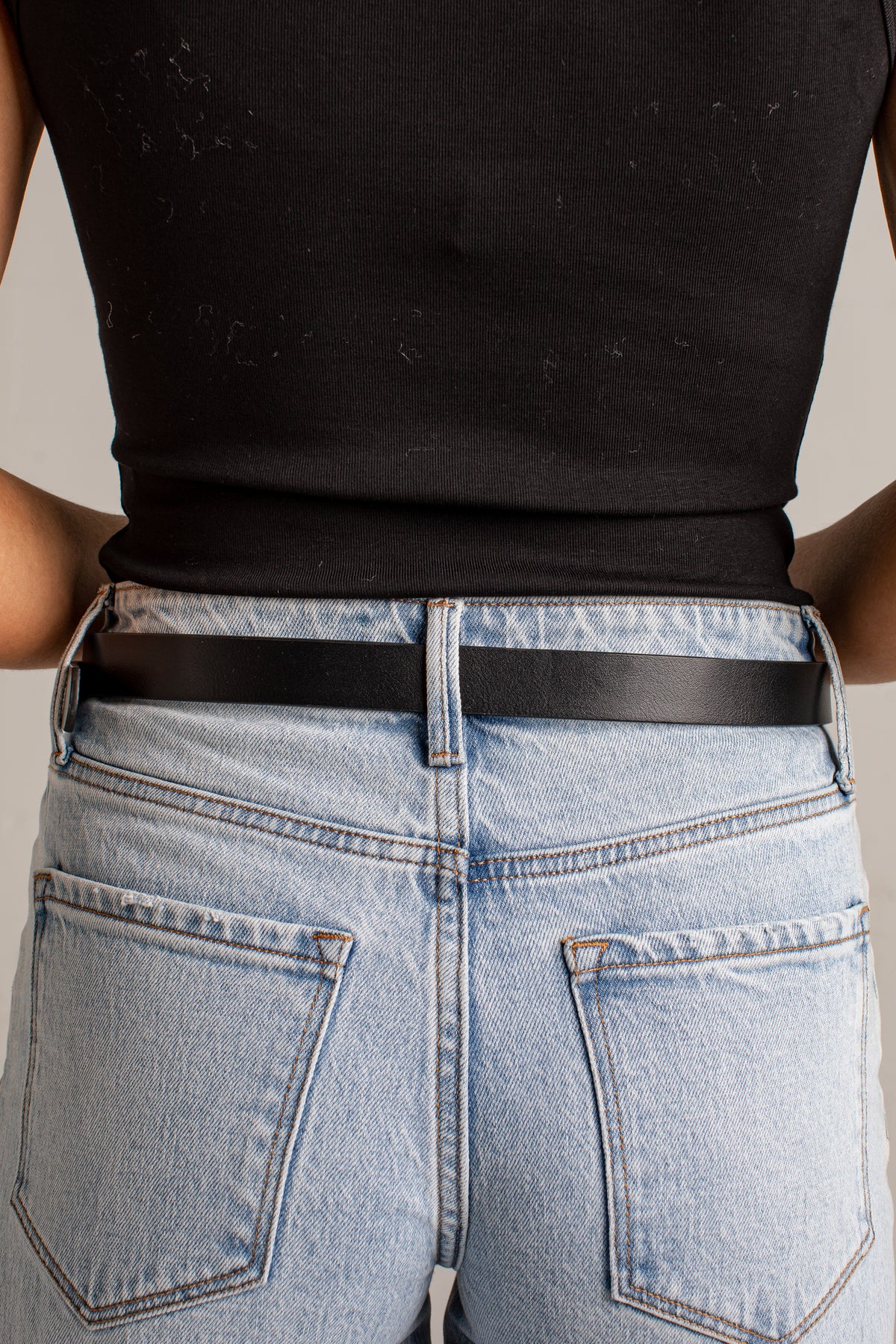 Close-up back view of a person wearing a black ribbed tank top, light-wash blue jeans, and a slim black belt against a plain light gray background.