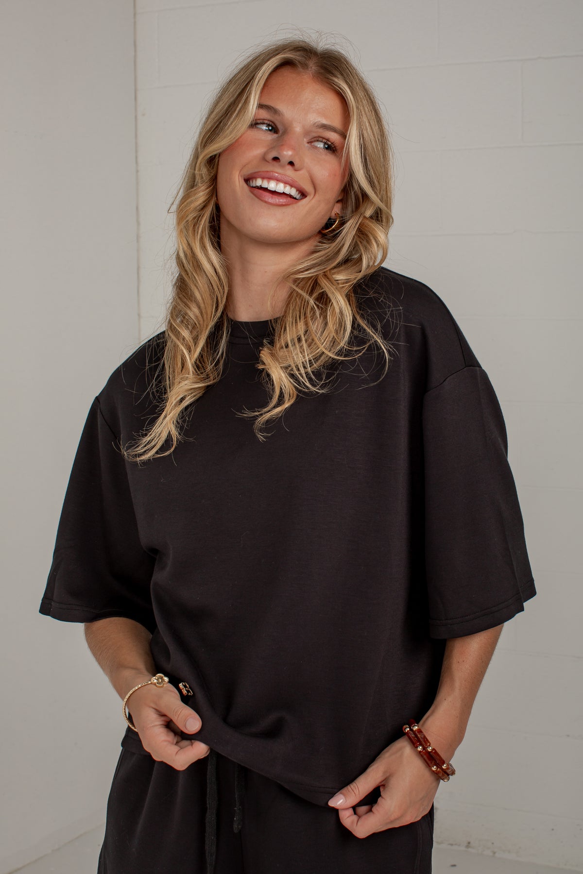 A smiling blonde woman wearing an oversized black short-sleeve t-shirt and matching black pants, standing against a white paneled wall.