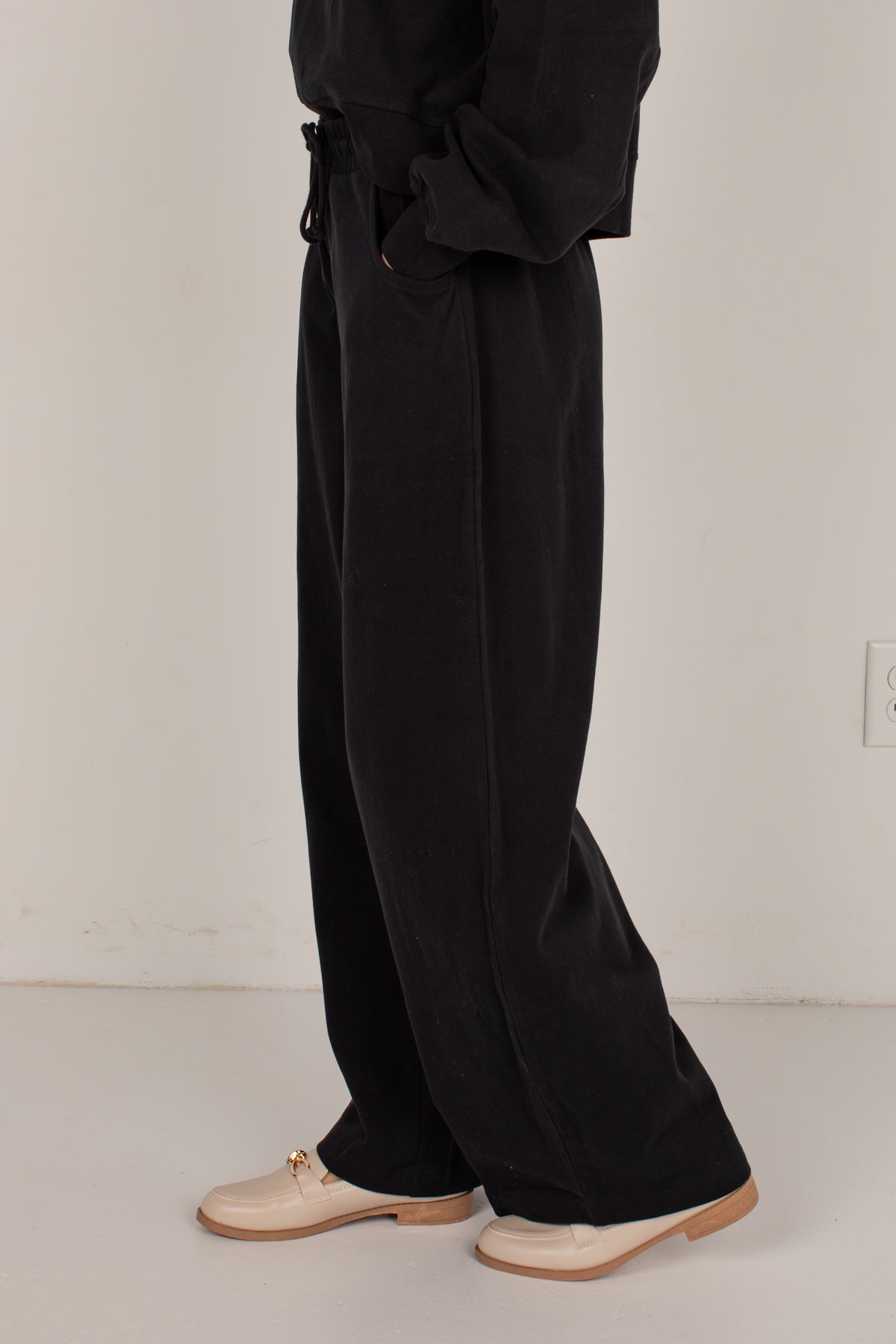 A person shown from the waist down wearing loose black wide-leg trousers, a black top, and beige loafers against a plain off-white wall.