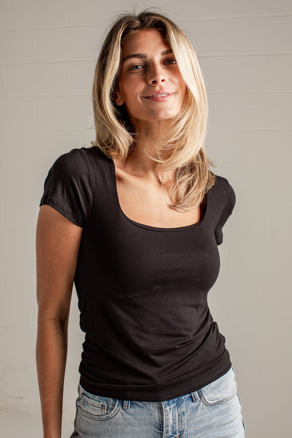 A woman with blonde hair wearing a black scoop-neck short-sleeve t-shirt and light blue jeans, standing against a light gray brick wall.