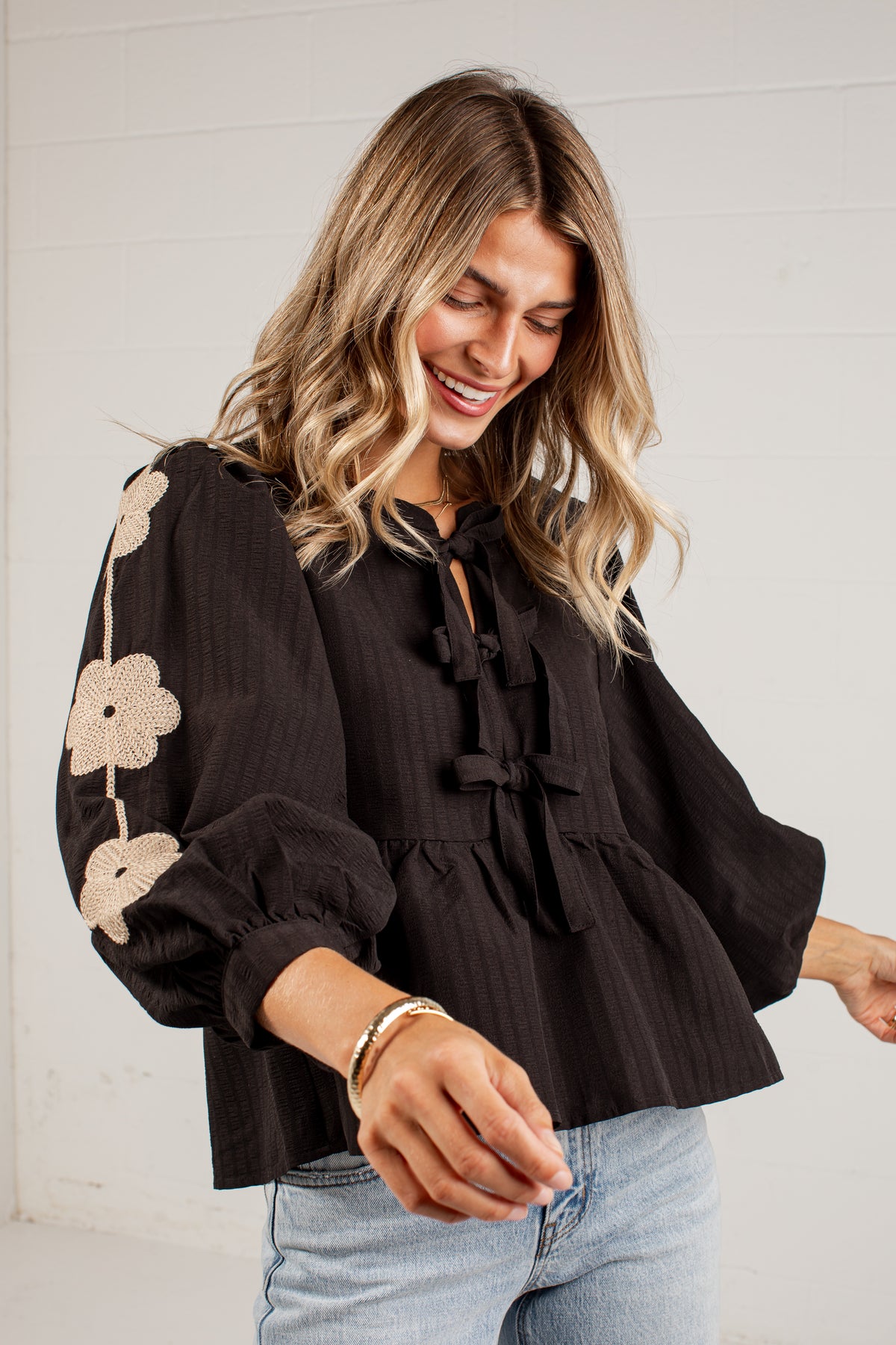 A woman wearing a black long-sleeve blouse with beige floral embroidery on the sleeve and tie-front bow details, paired with light blue jeans, standing against a white paneled wall.