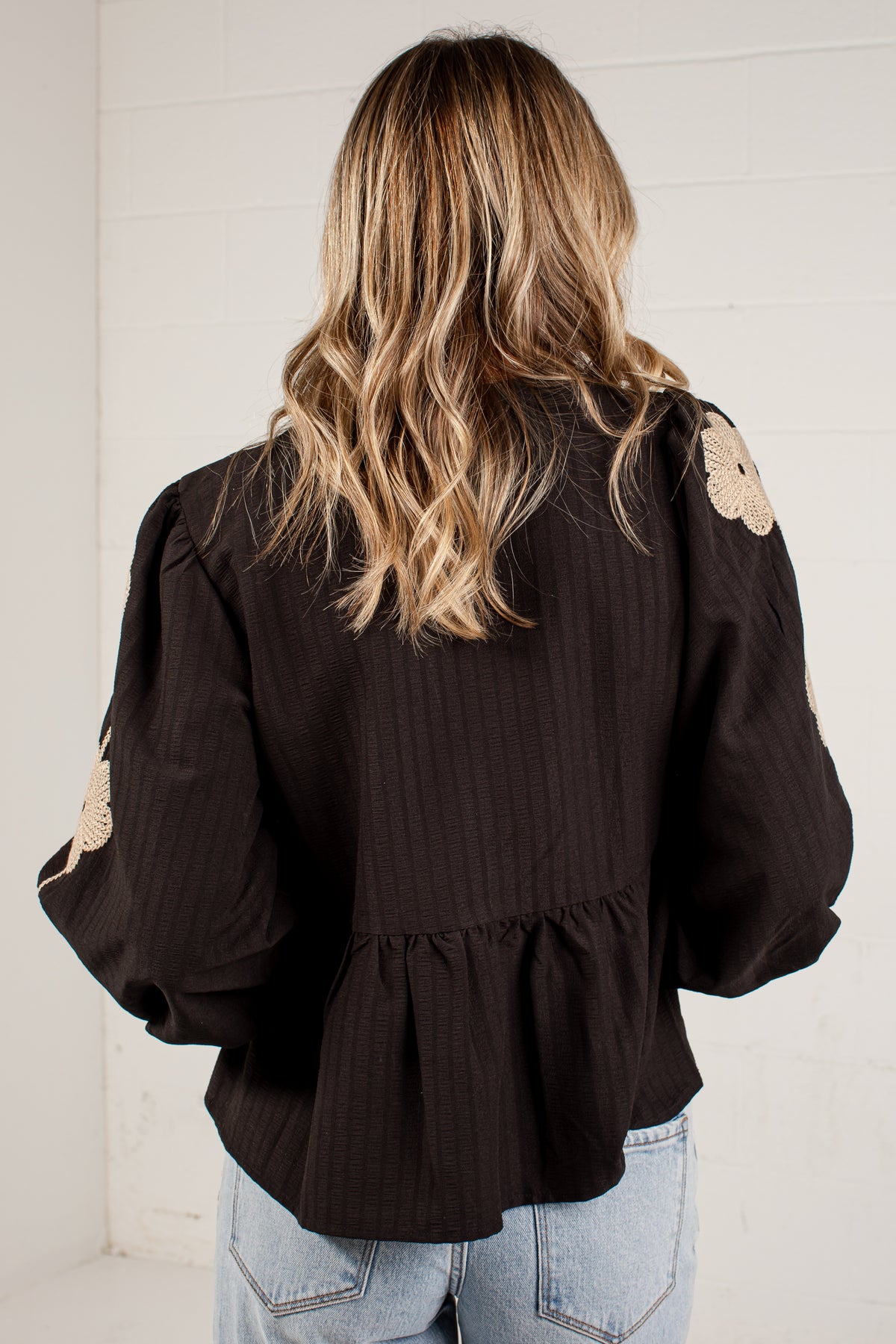 A woman viewed from behind wearing a dark brown long-sleeve blouse with beige floral appliques on the sleeves and a ruffled hem, paired with light blue jeans against a white paneled wall.
