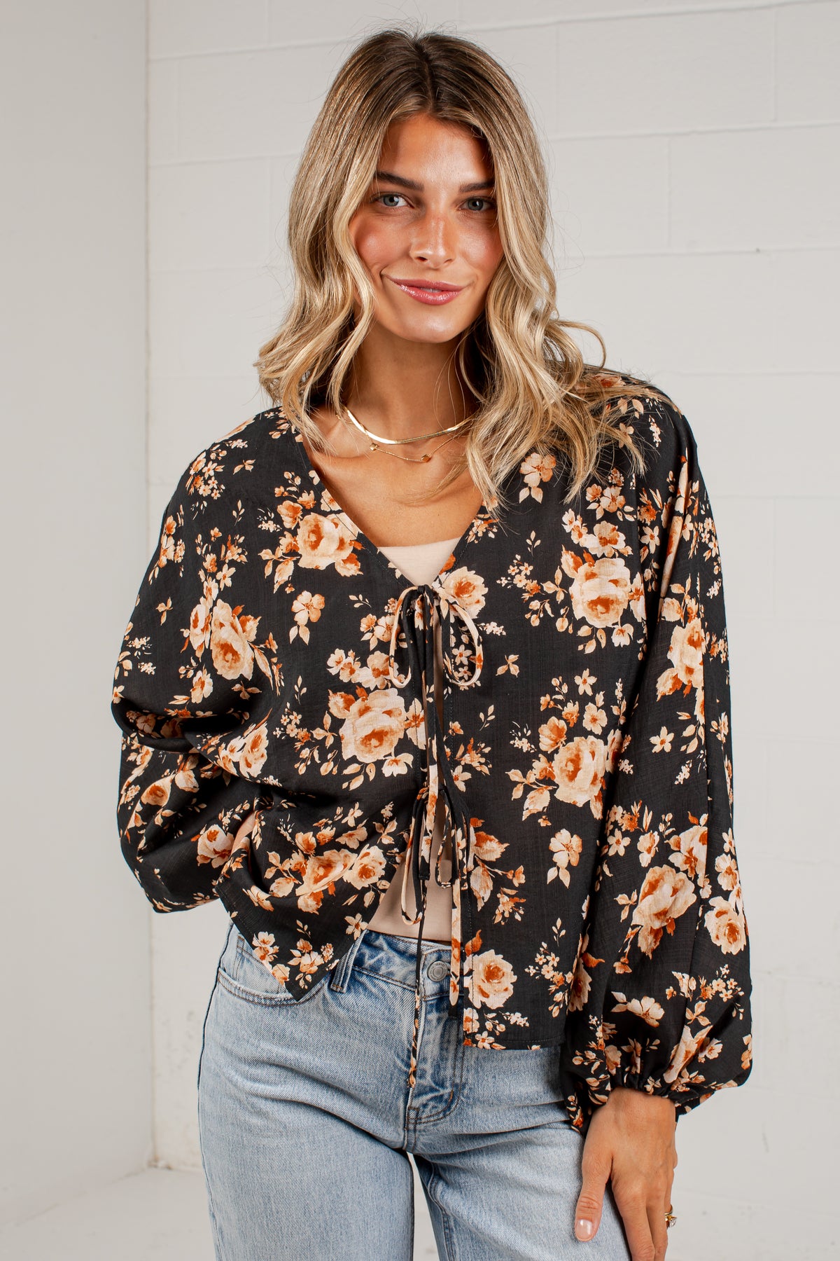 A woman wearing a black long-sleeve blouse with a warm-toned orange and beige floral print, featuring a tie-front detail and gathered sleeves, paired with light-wash blue jeans.