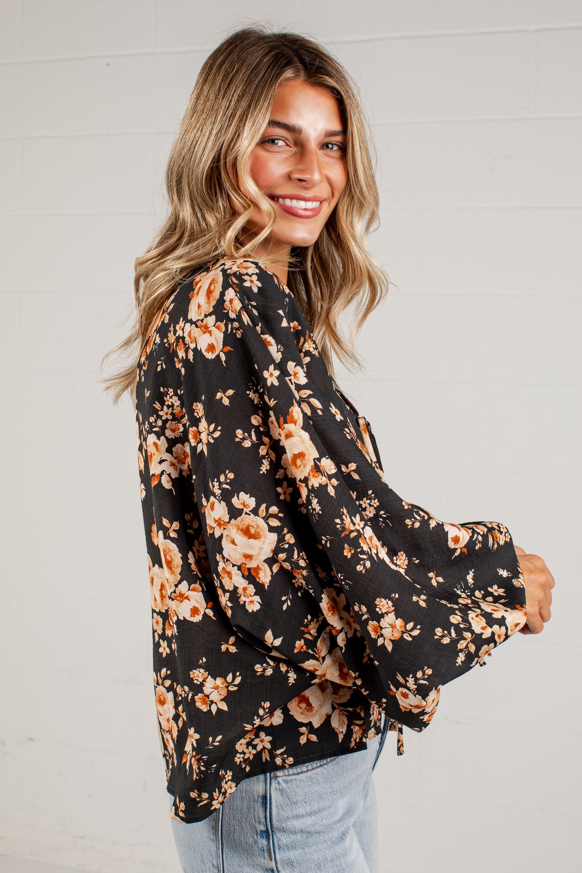 A woman wearing a black long-sleeve blouse with a warm-toned orange and beige floral pattern, paired with light blue jeans, standing against a white paneled wall.