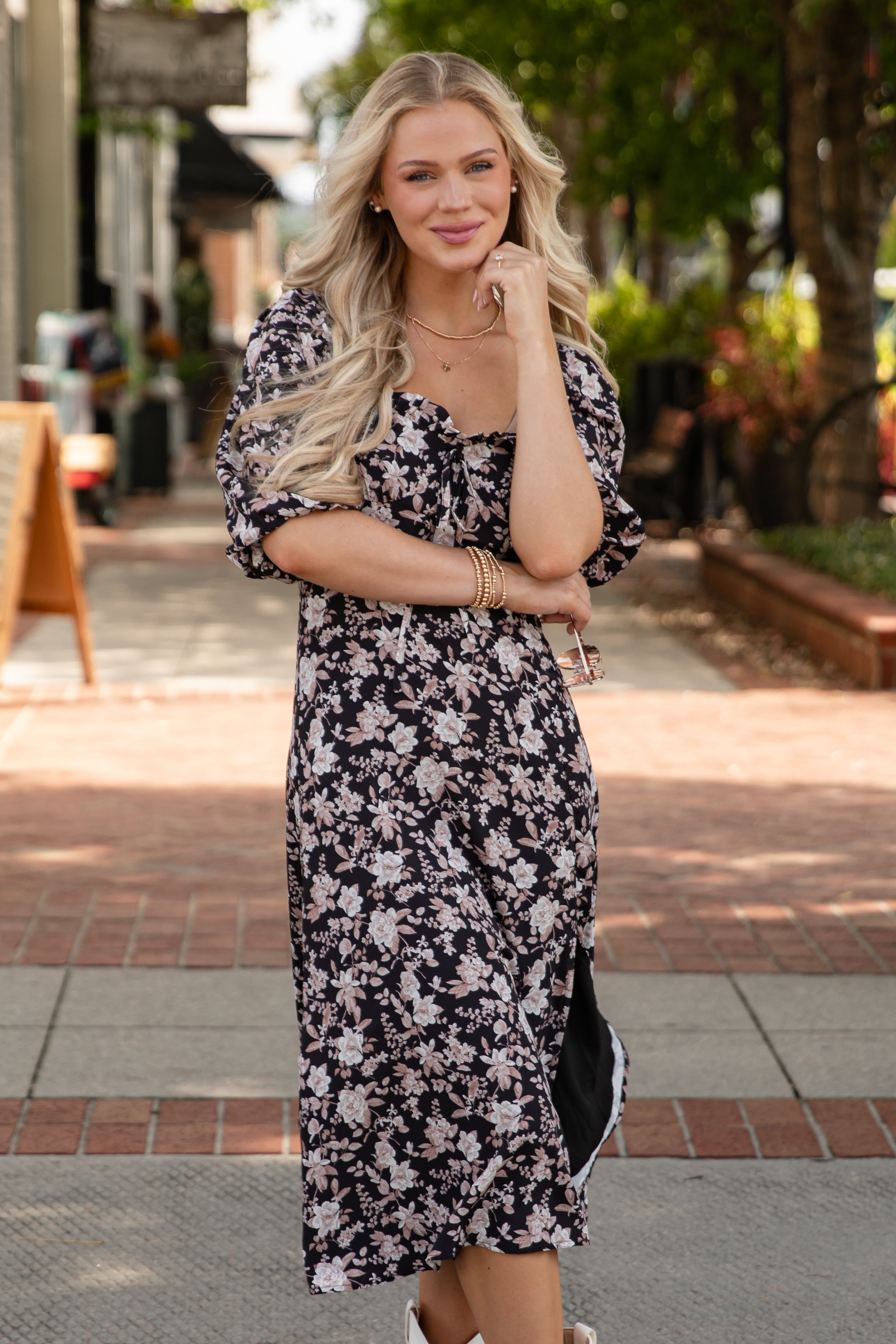 Woman in a floral dress standing on a sidewalk with trees and buildings in the background