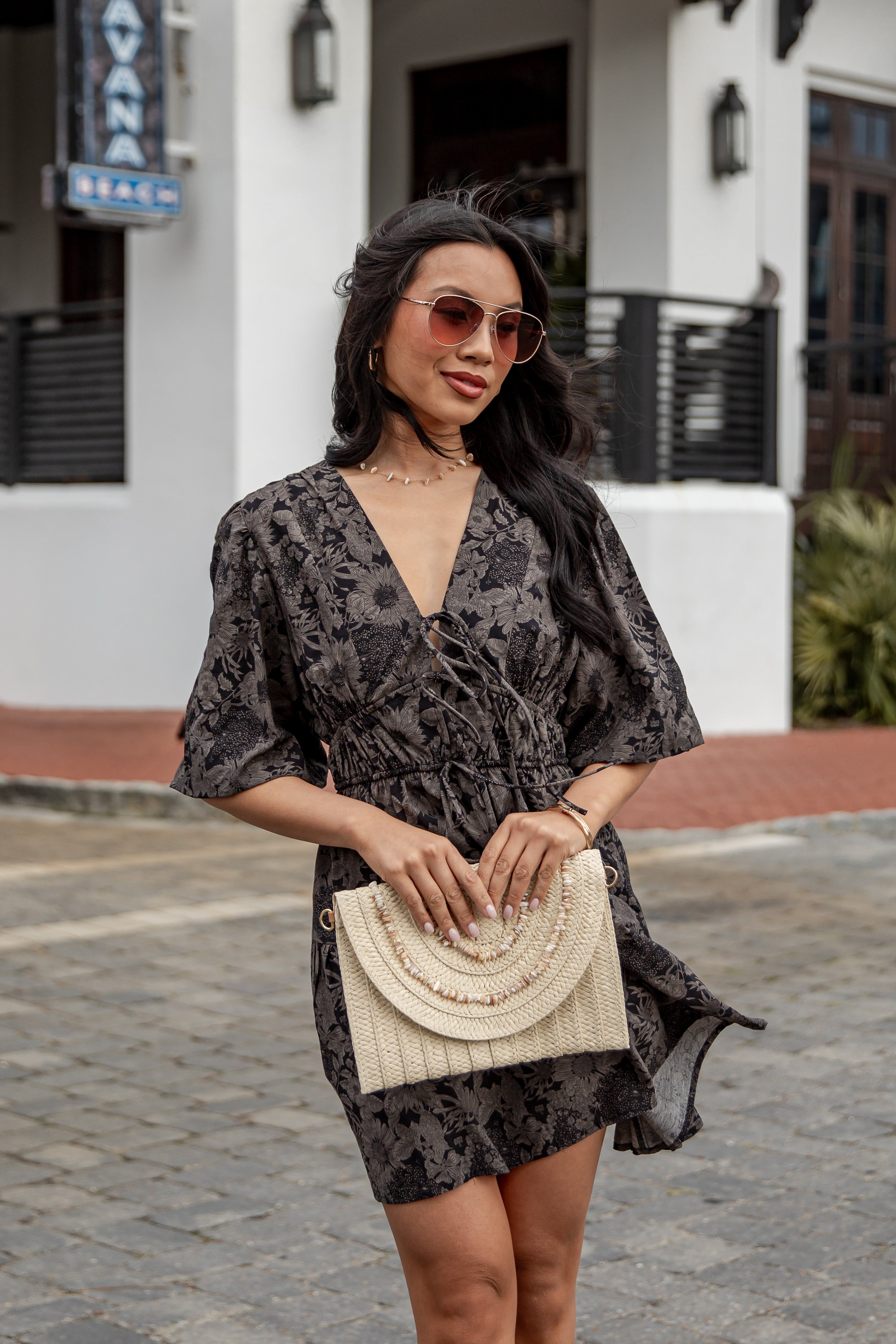 Woman in a black floral dress holding a beige handbag on a street.