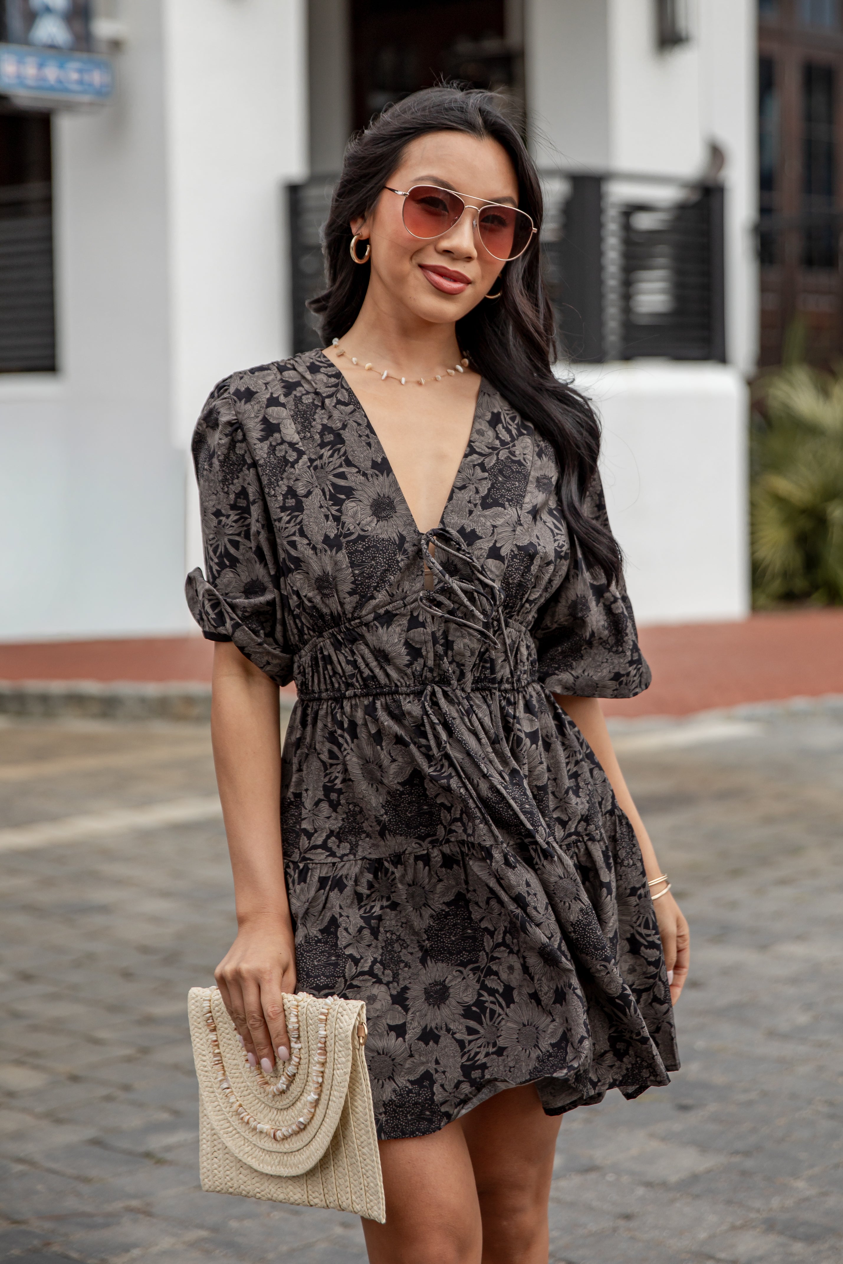 Woman wearing a black floral dress with sunglasses and a beige clutch in an urban setting.