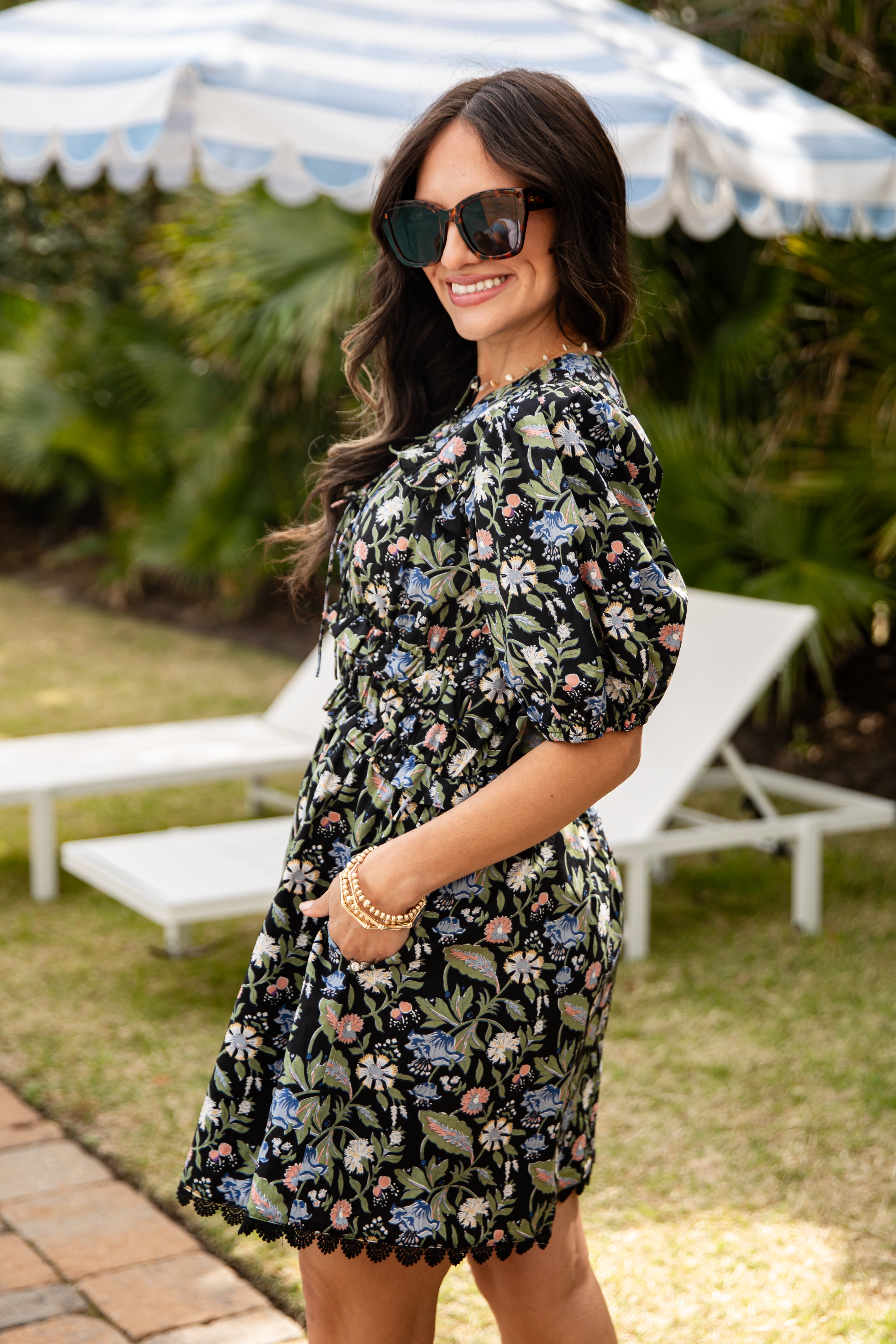 Woman wearing a floral dress standing outdoors with lounge chairs and an umbrella in the background.