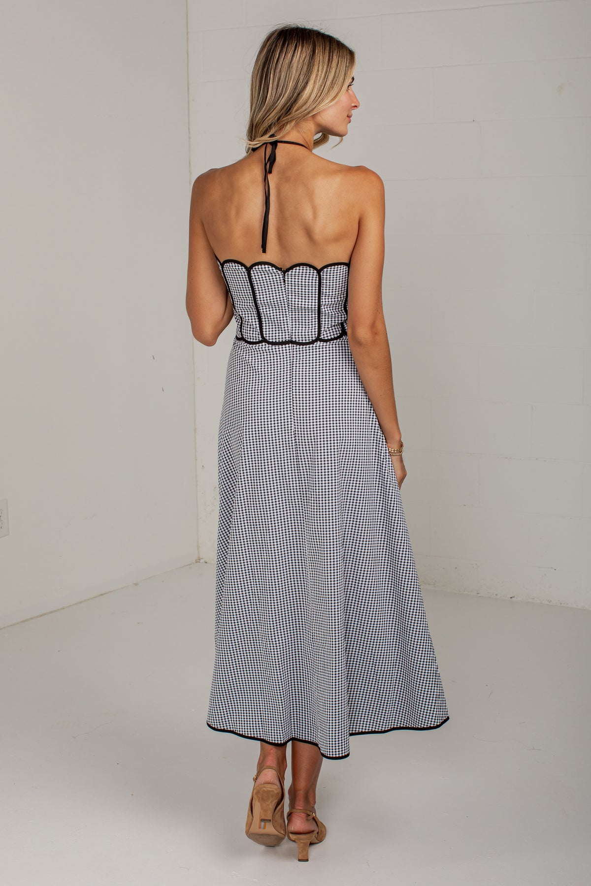 A woman shown from the back wearing a sleeveless midi dress with thin vertical black and white stripes, a scalloped black trim at the waist, and a halter-style back strap, paired with tan heeled sandals in a white studio setting.