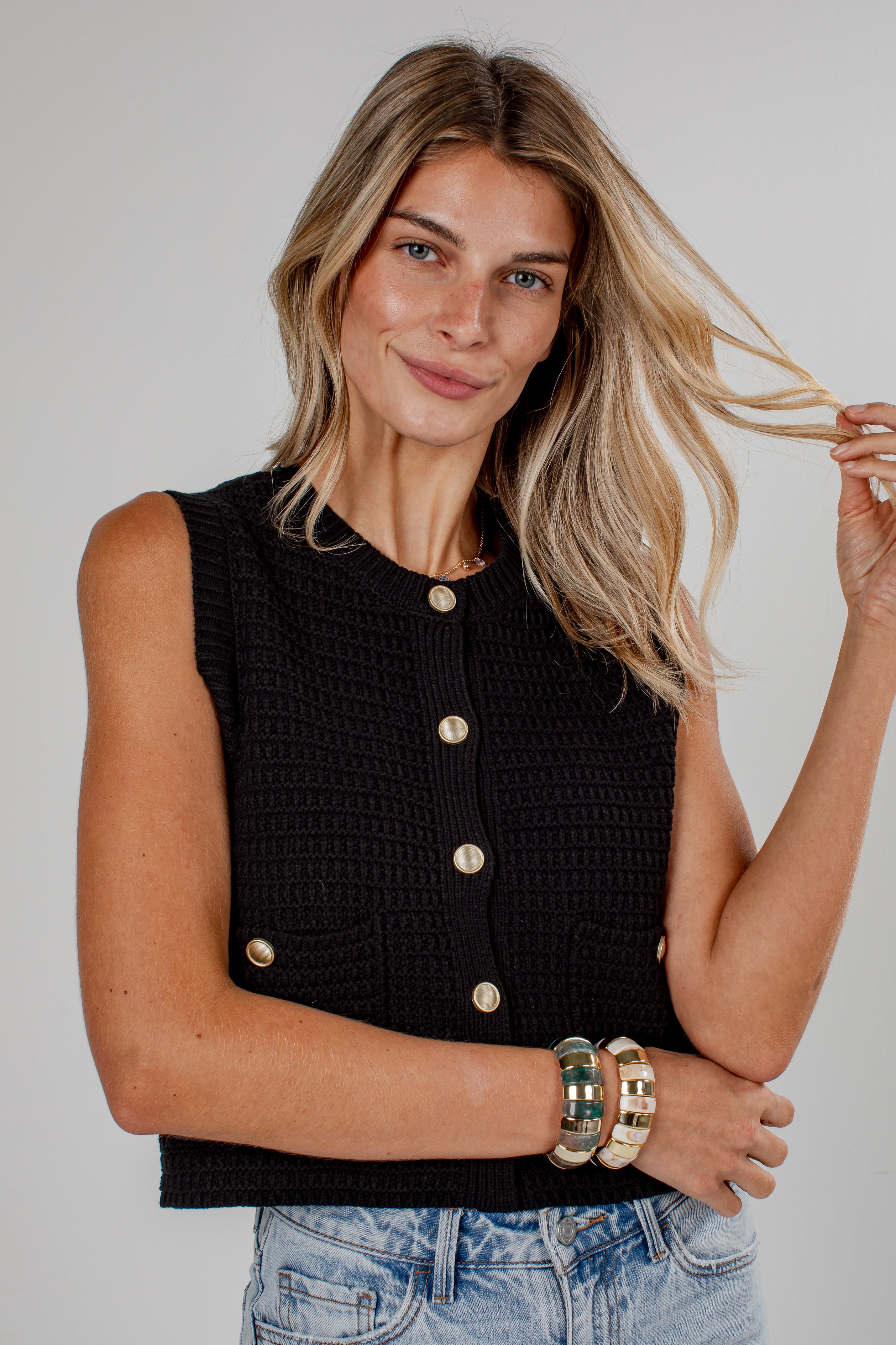 A woman wearing a black sleeveless textured knit top with gold buttons, paired with light blue jeans, against a plain light gray studio background.