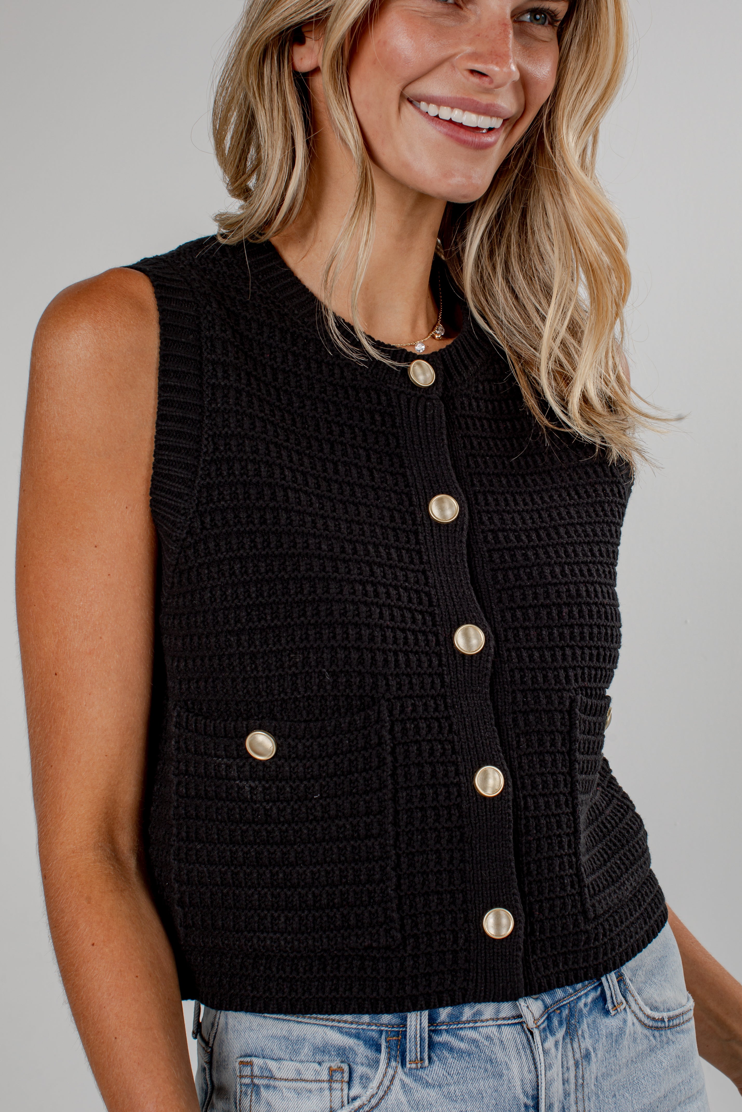 A woman wearing a sleeveless black knit vest with gold-tone buttons and front pockets, paired with light blue jeans, against a plain light gray background.