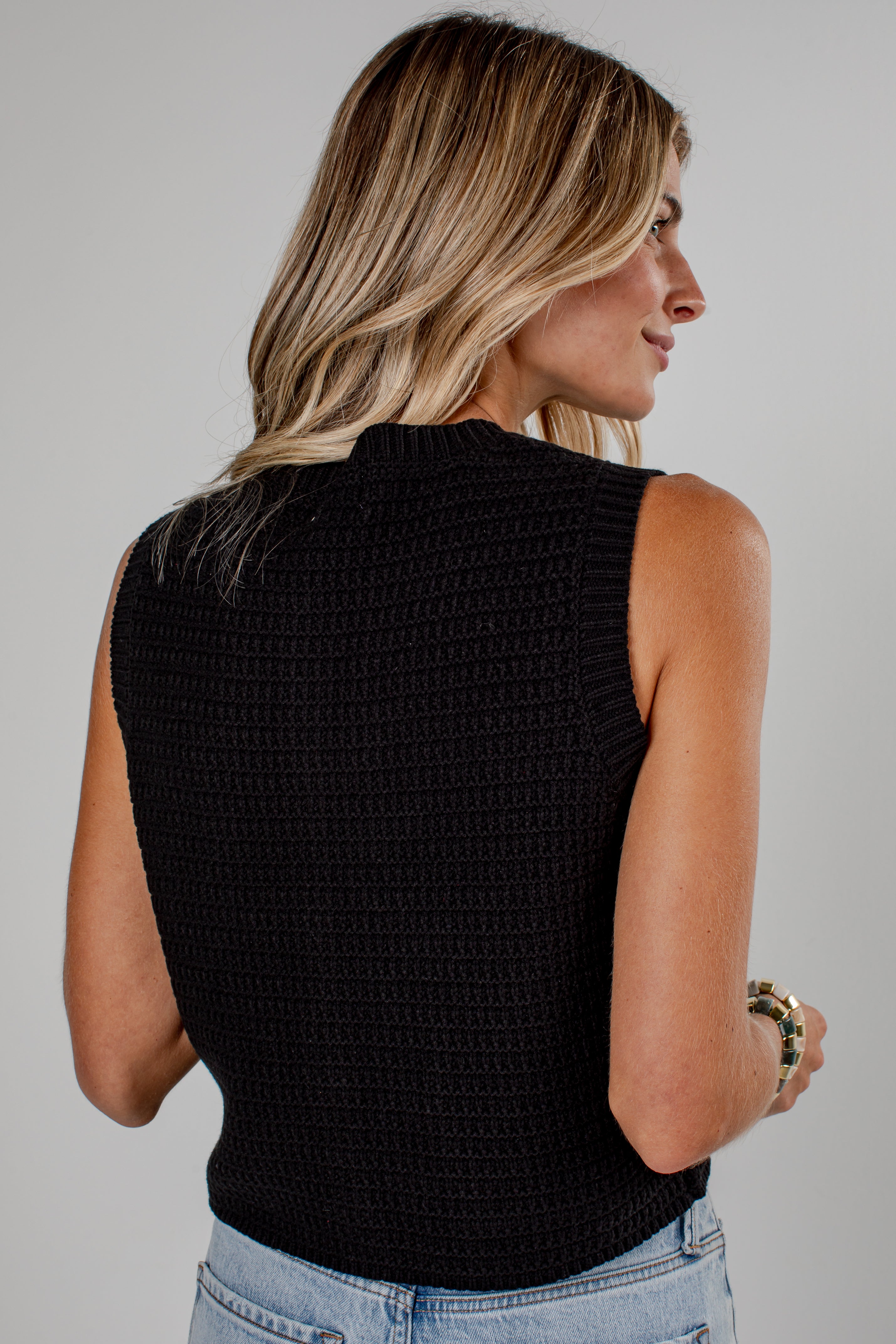 A woman shown from the back wearing a black sleeveless knit top with a textured waffle pattern, paired with light blue jeans, against a plain light gray background.