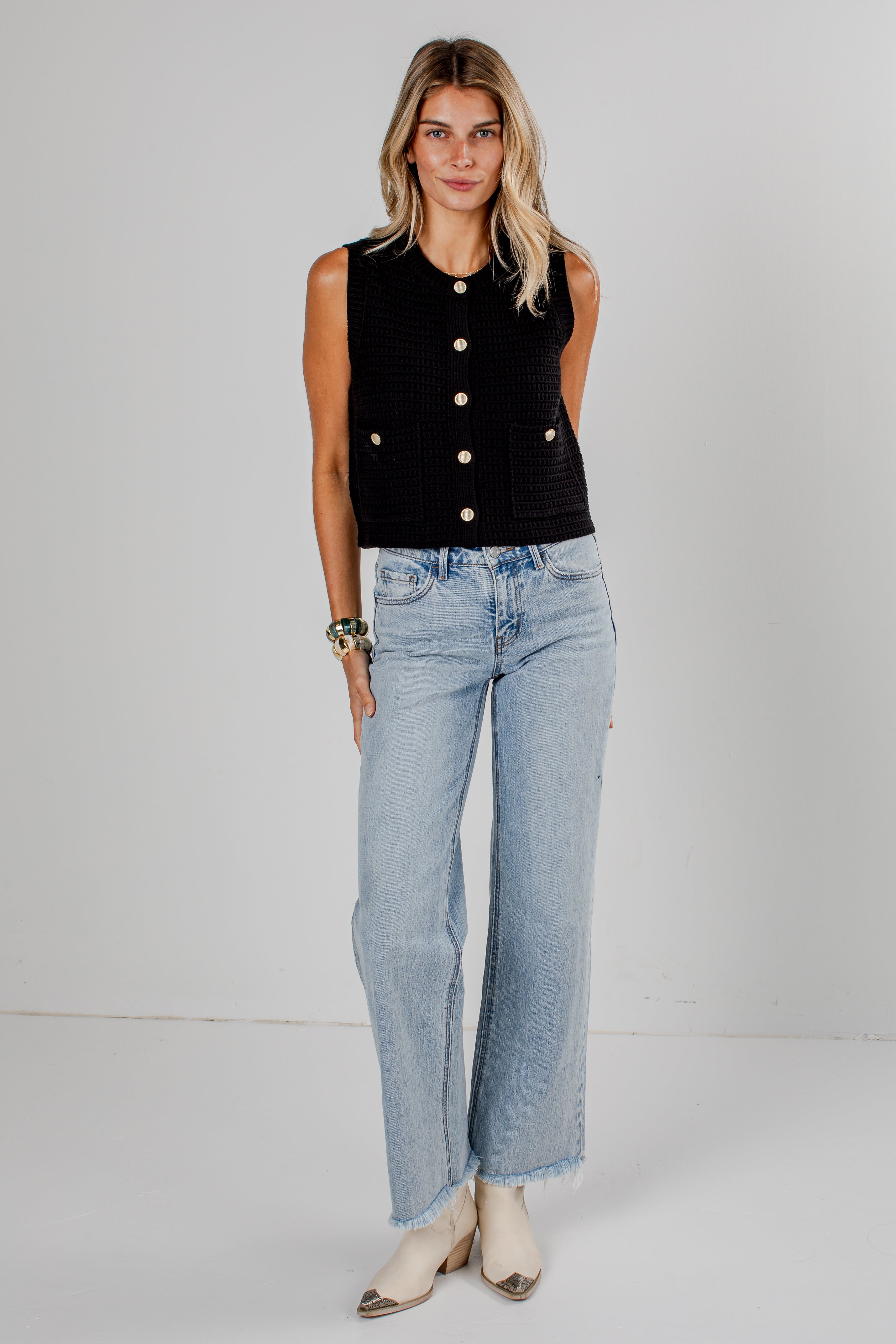 A woman modeling a sleeveless black textured vest with gold buttons and light-wash wide-leg jeans, standing against a plain light gray studio background.