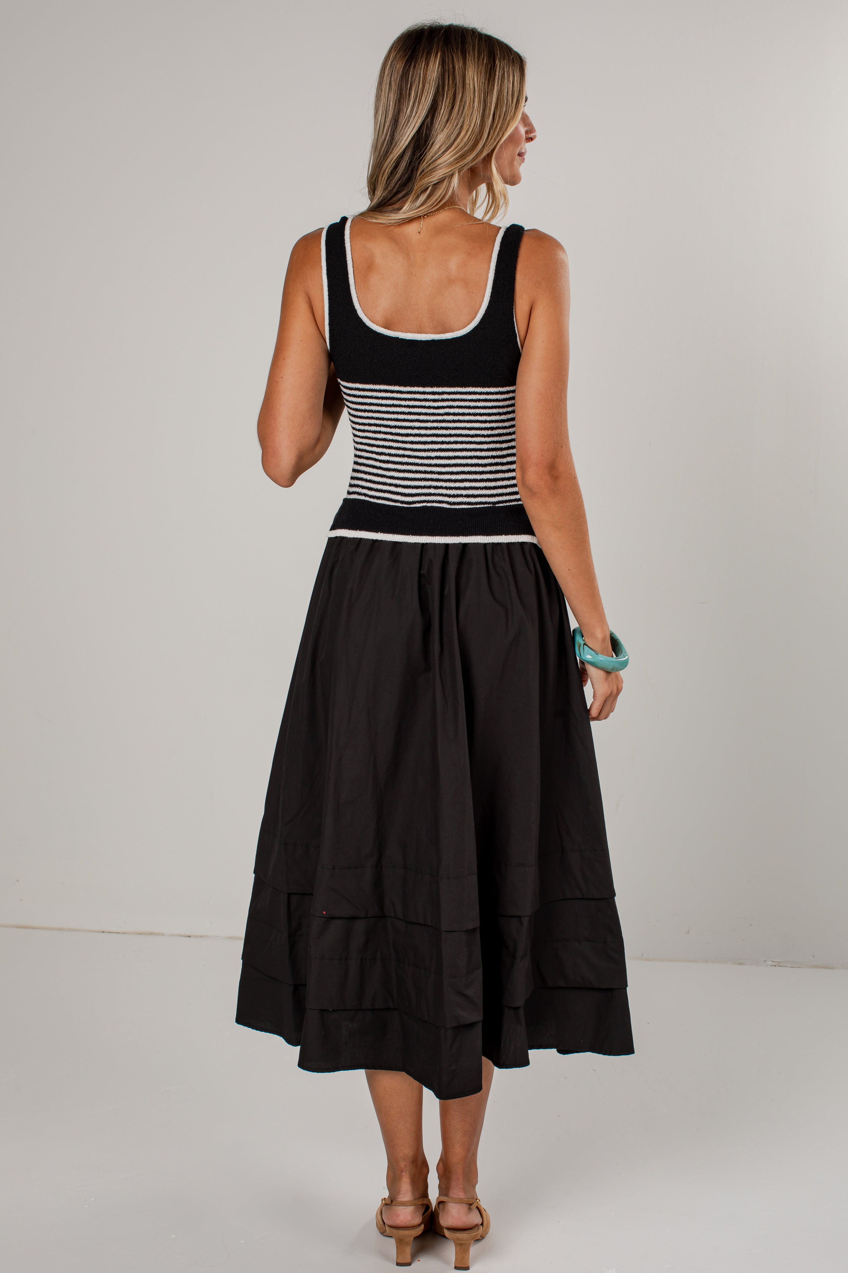 A woman shown from the back wearing a sleeveless black midi dress with a black and white horizontally striped bodice and a full black skirt, photographed against a plain light gray studio background.