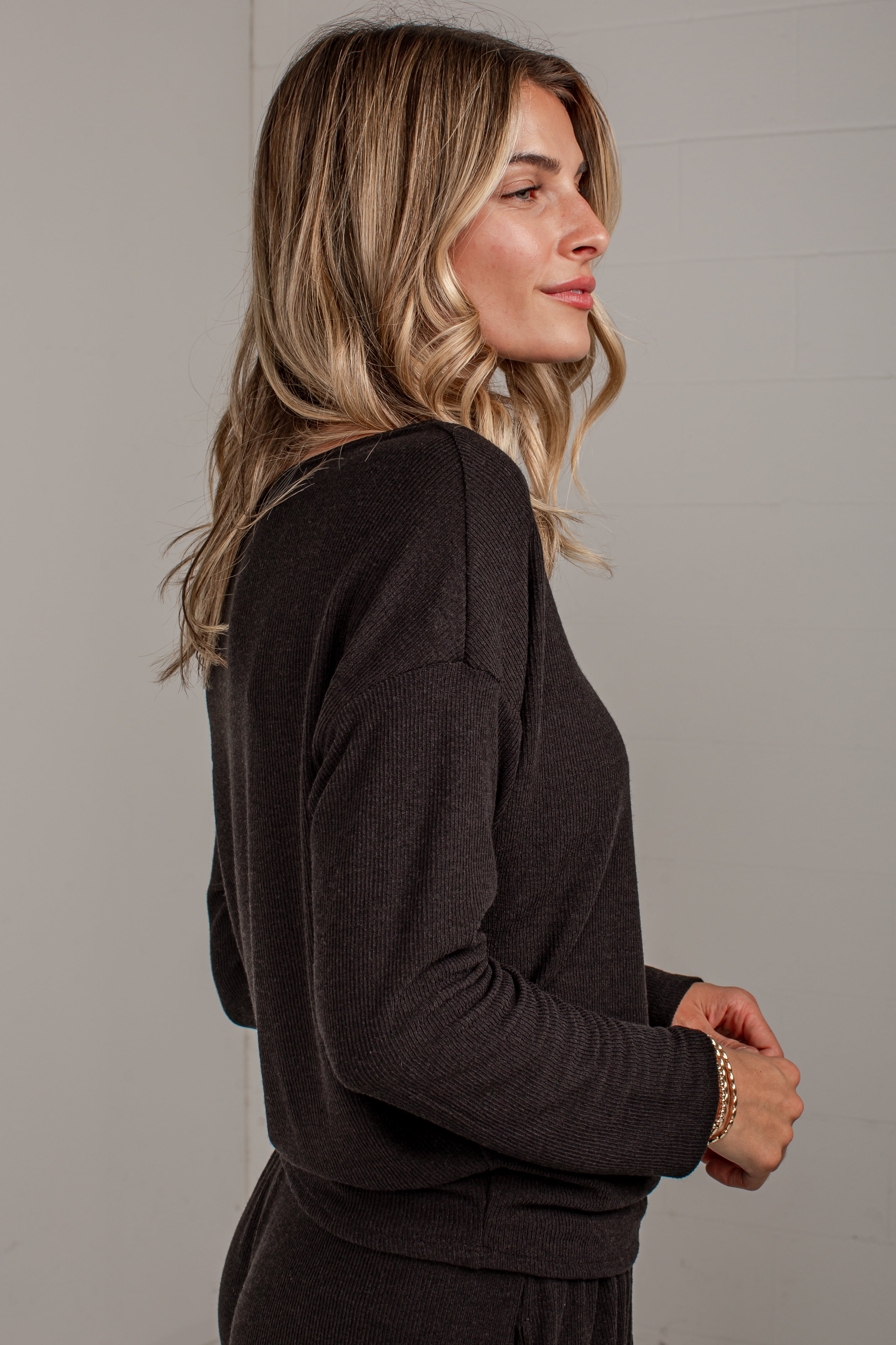 A woman with blonde-highlighted hair is shown in profile wearing a dark brown long-sleeve knit top and matching bottoms against a light gray wall.