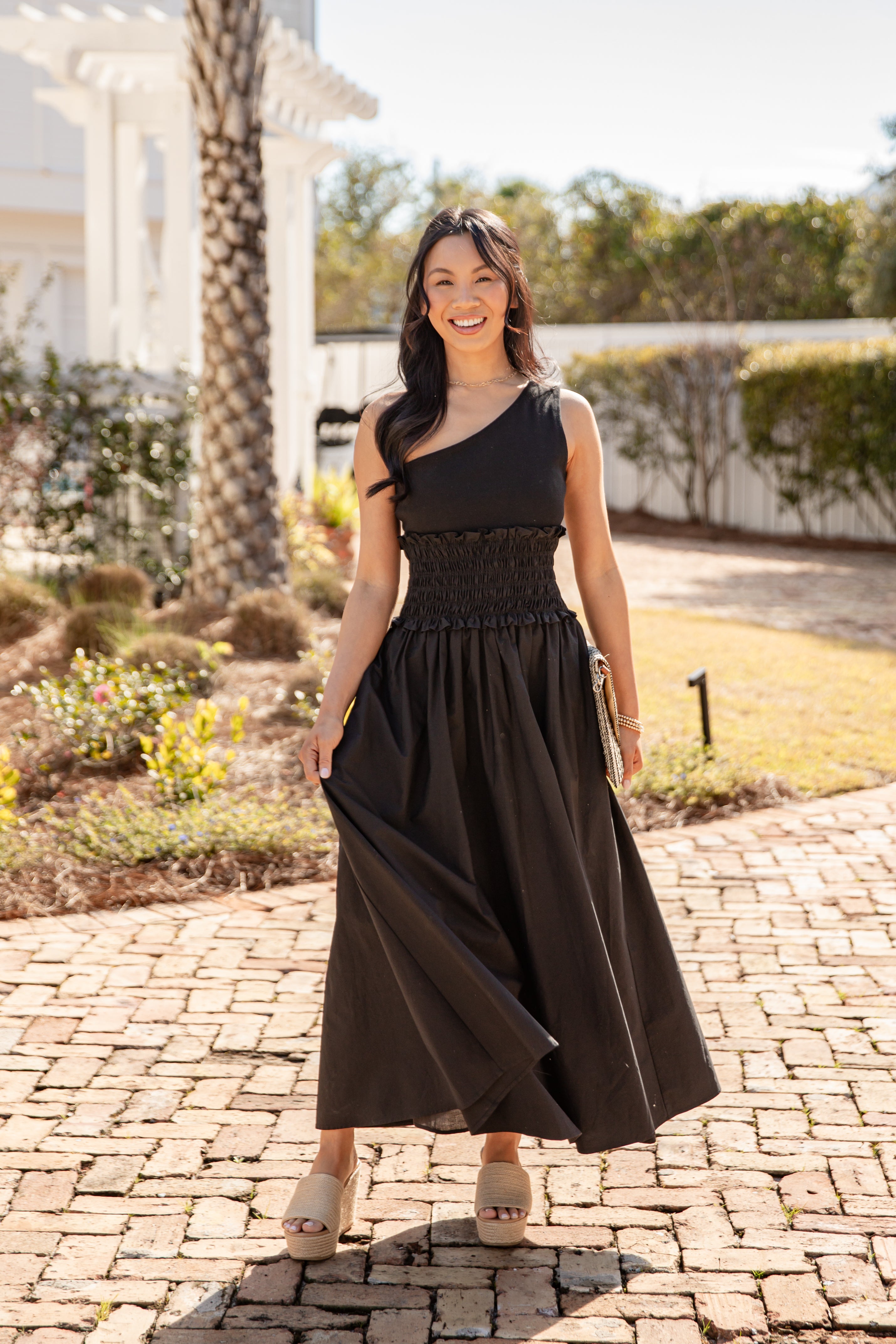 Woman in a black one-shoulder dress standing on a paved walkway with greenery in the background