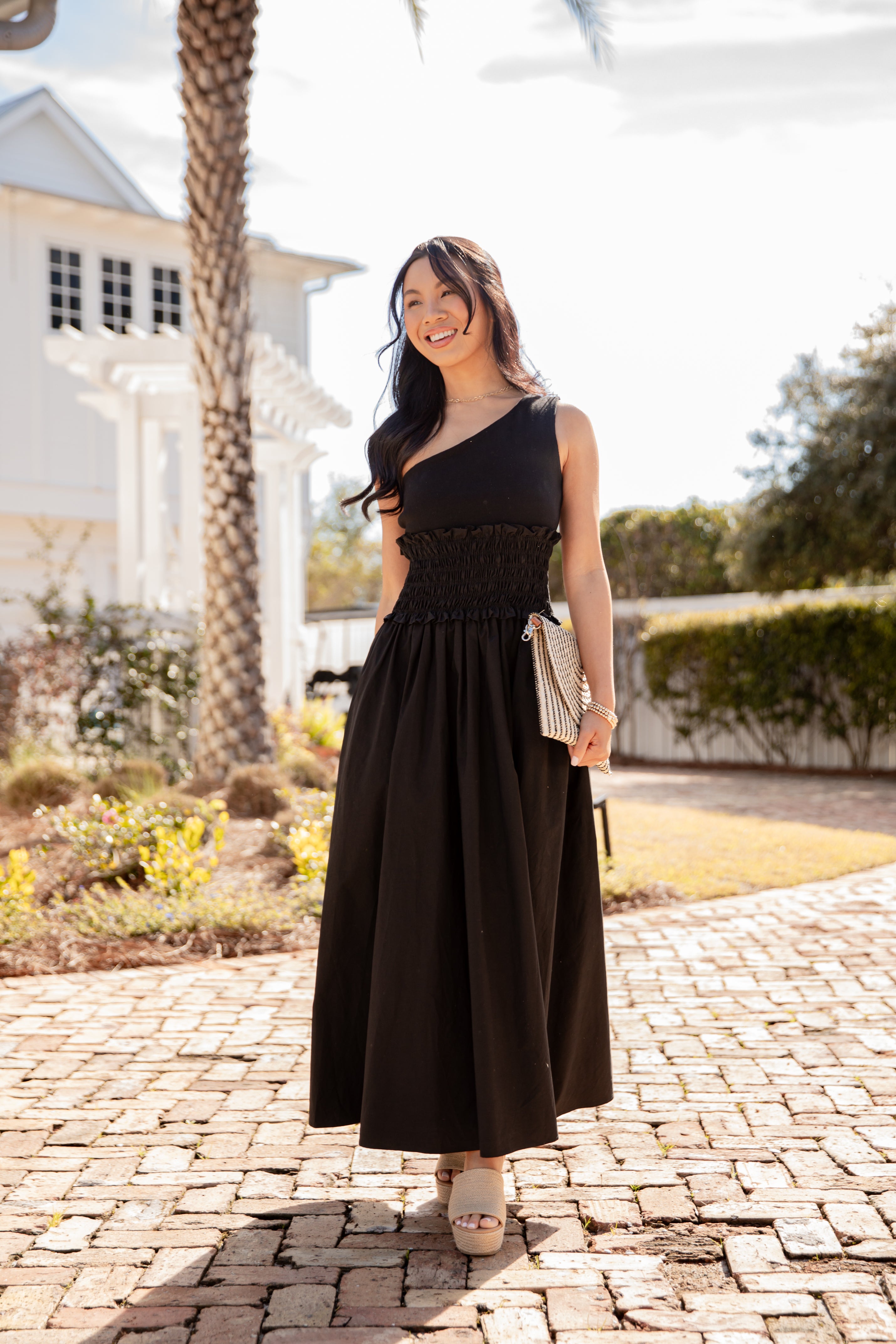 Woman in a black dress standing on a brick path with a house and palm tree in the background.