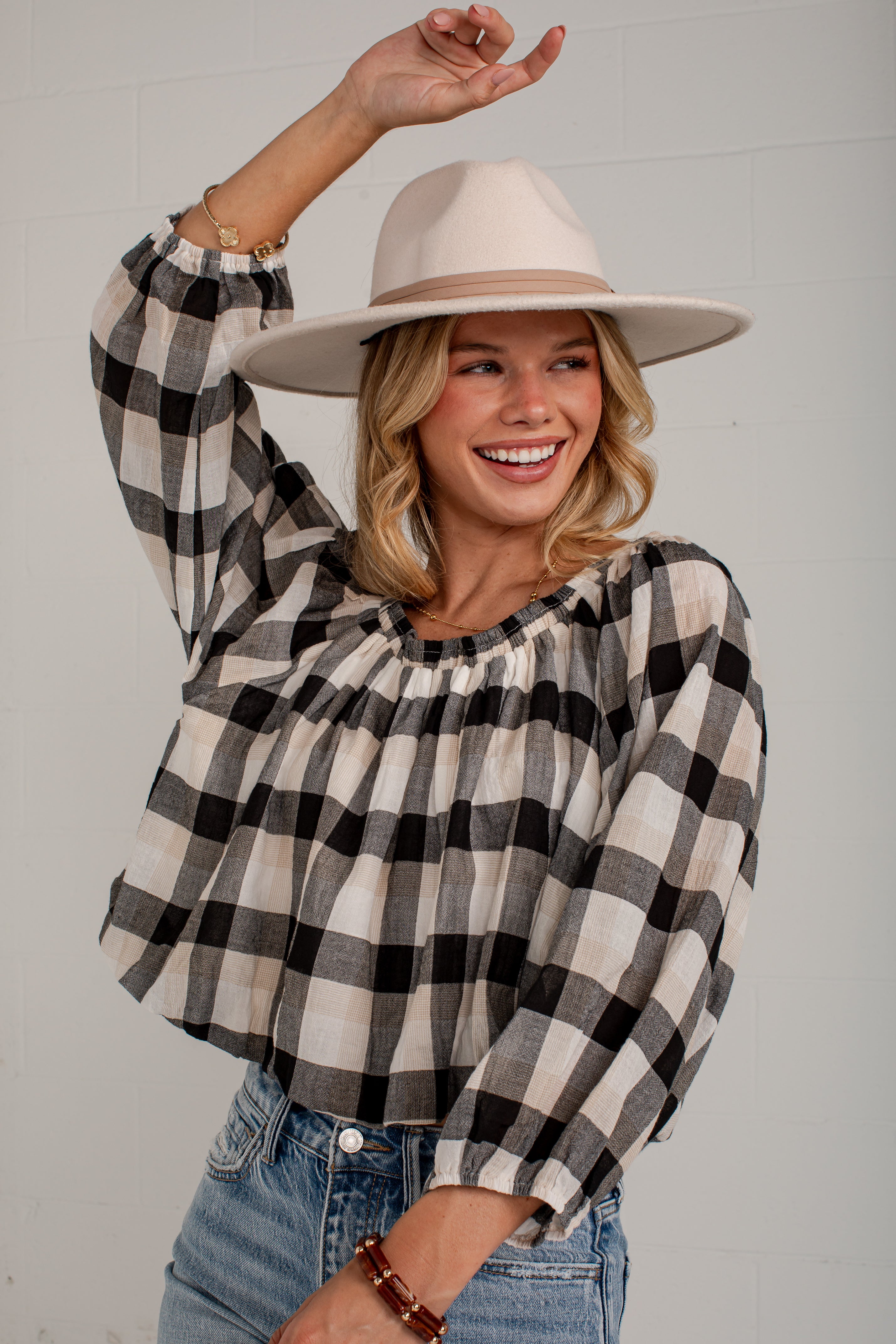 A woman wearing a black and beige buffalo plaid long-sleeve blouse with a gathered neckline, paired with a beige wide-brim hat and blue jeans, posing against a white paneled wall.