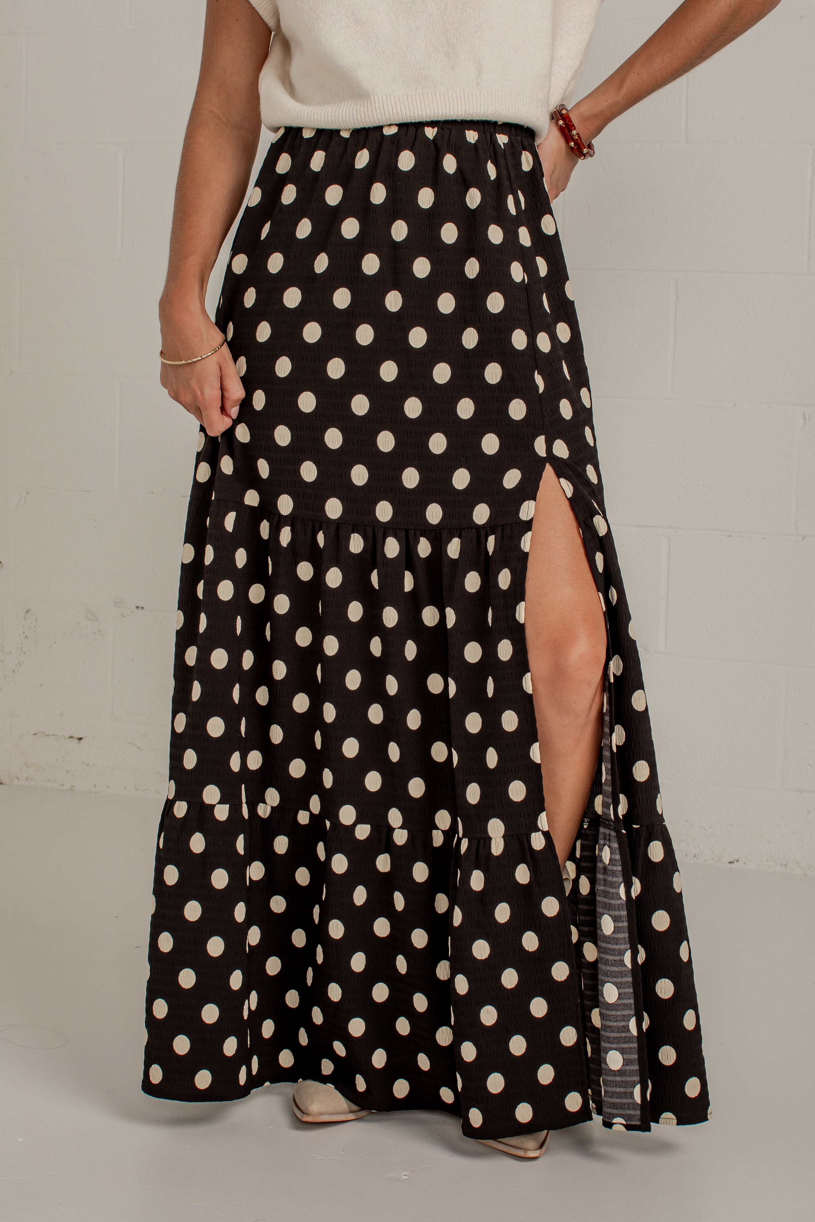 A woman wearing a long black maxi skirt with a cream polka dot pattern and a high side slit, paired with a cream top against a light gray wall.