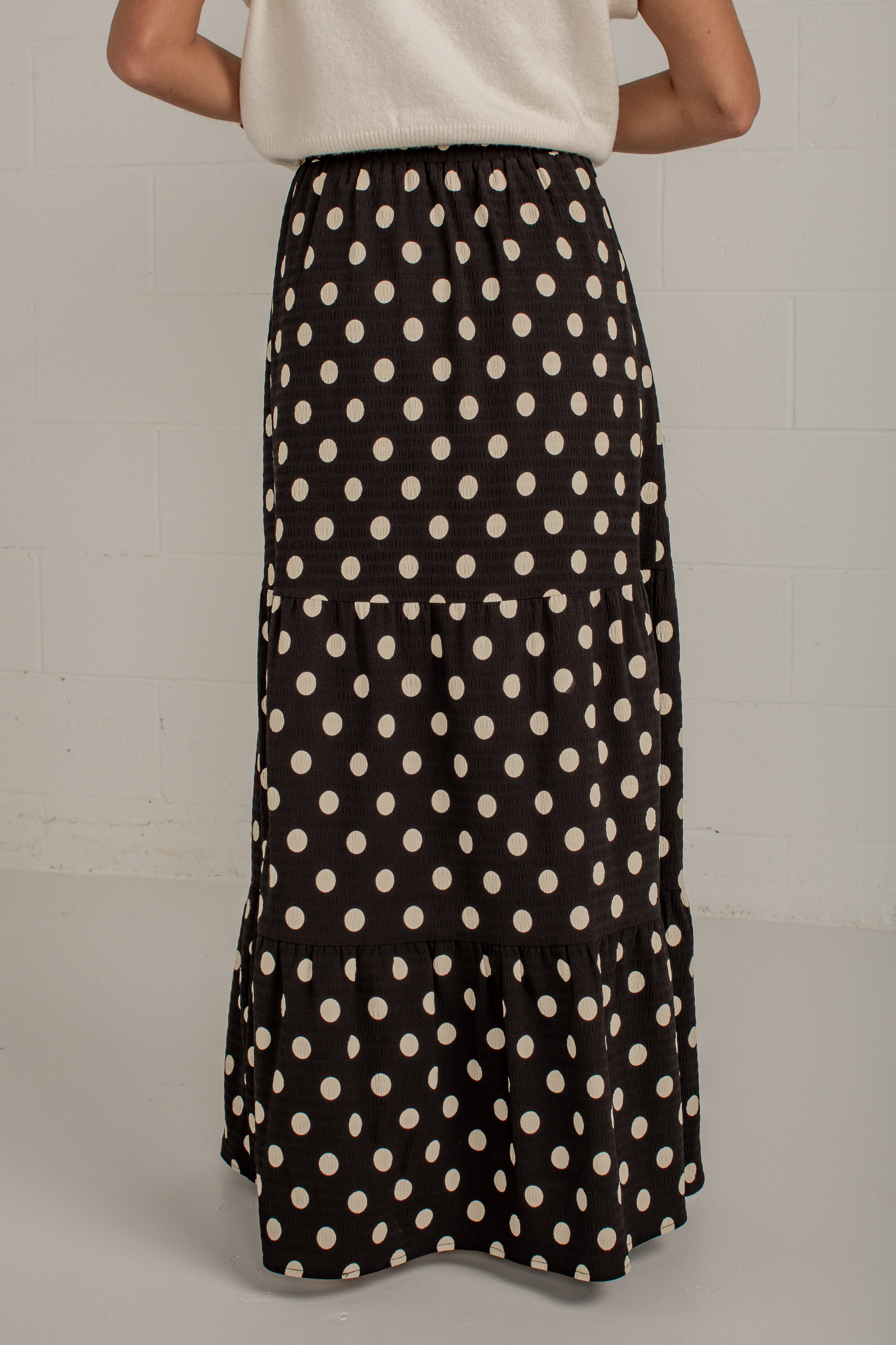A black tiered maxi skirt with a cream polka dot pattern, shown on a model from the back against a light gray brick wall.