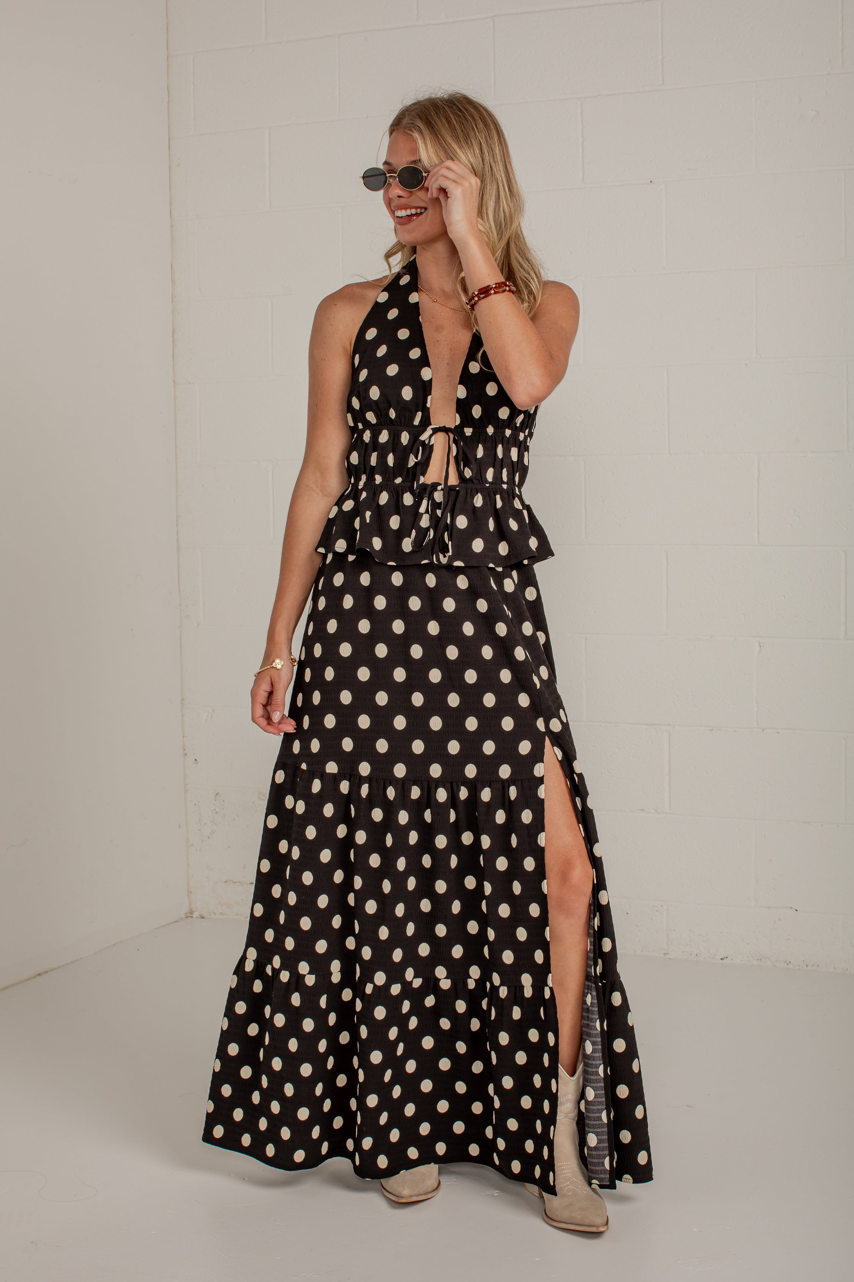 A woman modeling a sleeveless black halter maxi dress with a beige and white polka dot pattern, featuring a tie-front waist and a high side slit, set against a white brick wall.