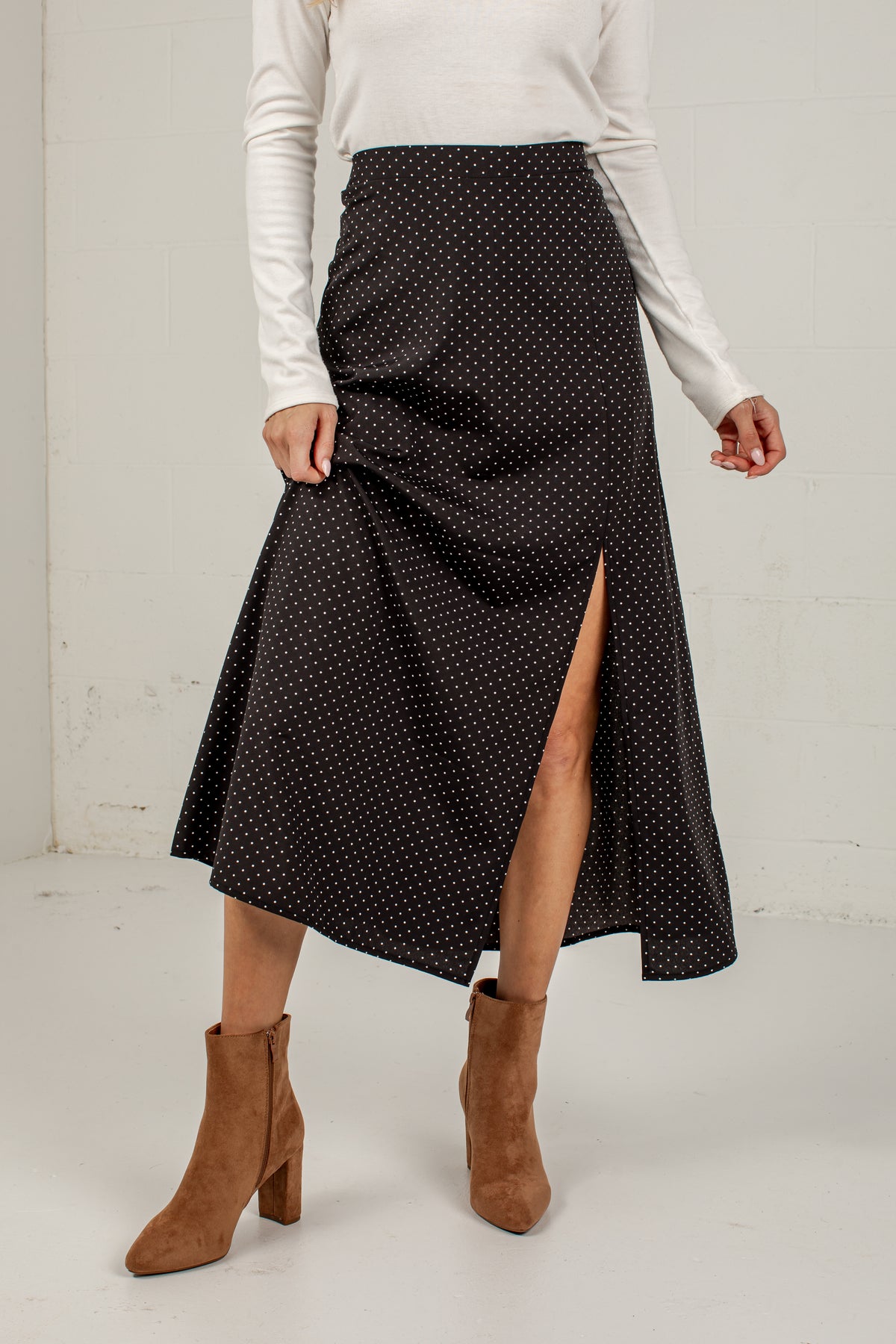 A woman wearing a black midi skirt with a small white polka dot pattern and a front slit, styled with a white long-sleeve top and brown suede ankle boots.