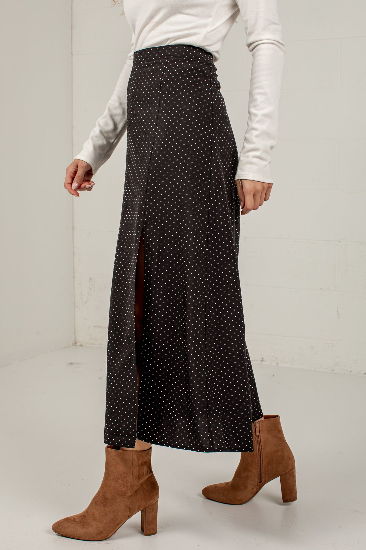 A model wearing a black midi skirt with a white polka dot pattern and a front slit, styled with a white long-sleeve top and brown suede ankle boots.