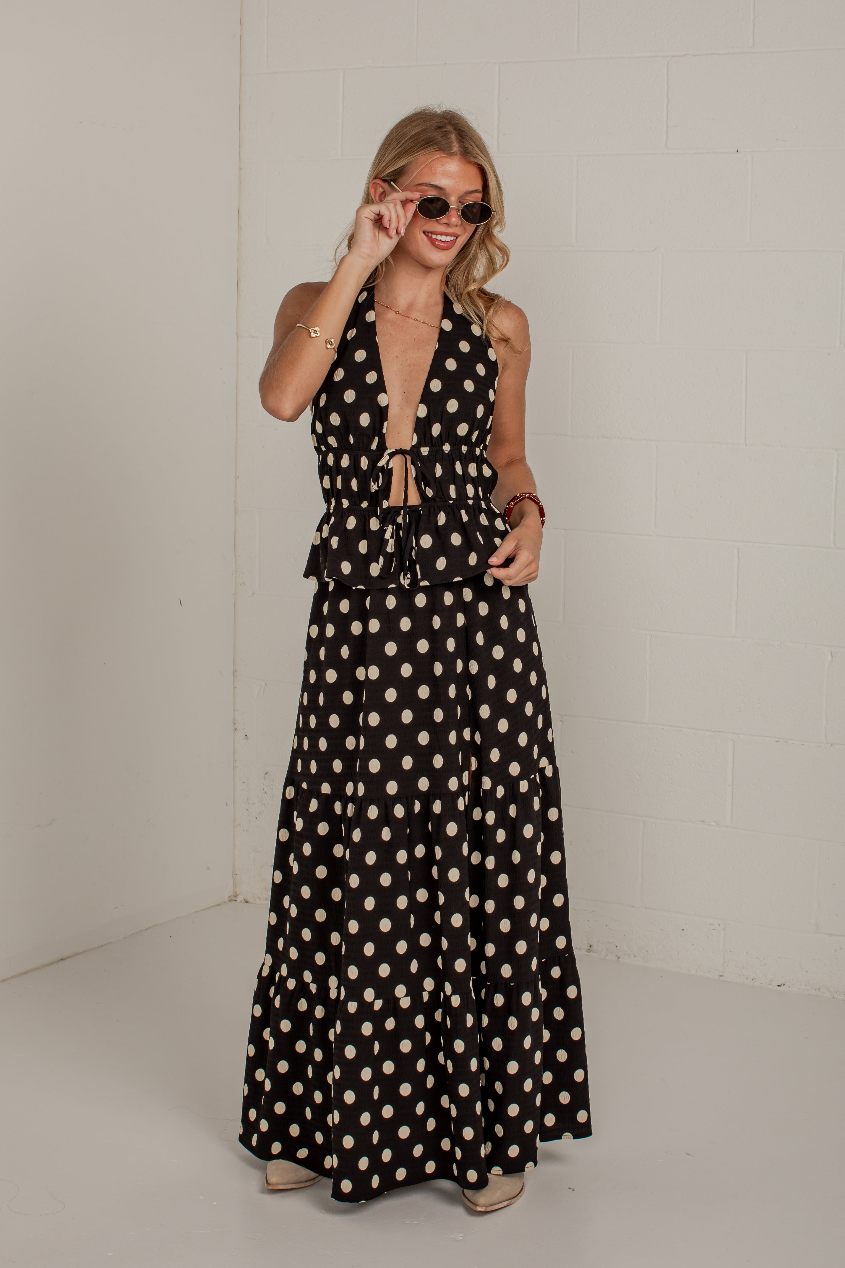 A woman modeling a black halter-neck maxi dress with a beige polka dot pattern, standing against a white brick wall.