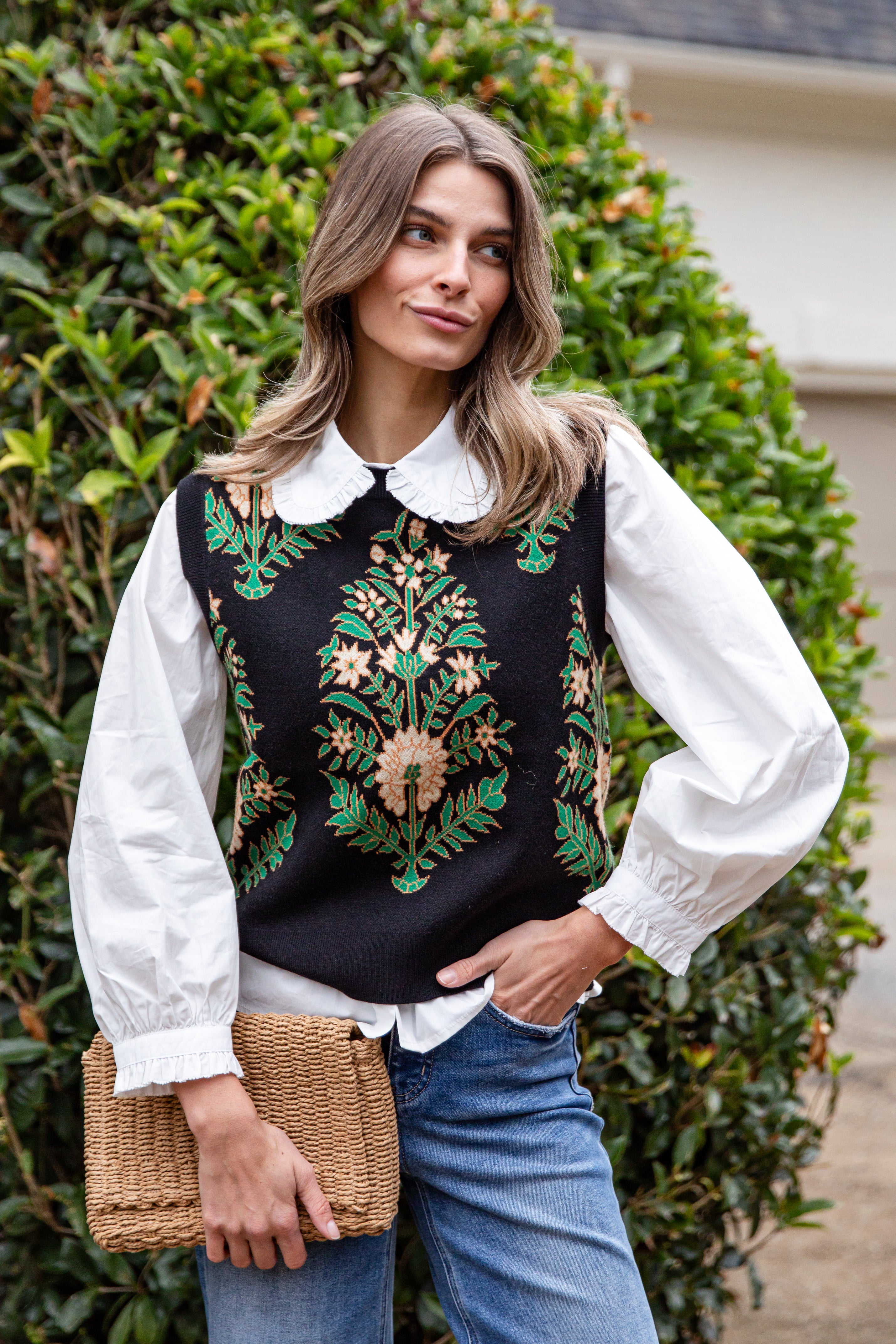Woman wearing a black embroidered vest over a white blouse with blue jeans, standing in front of green foliage.