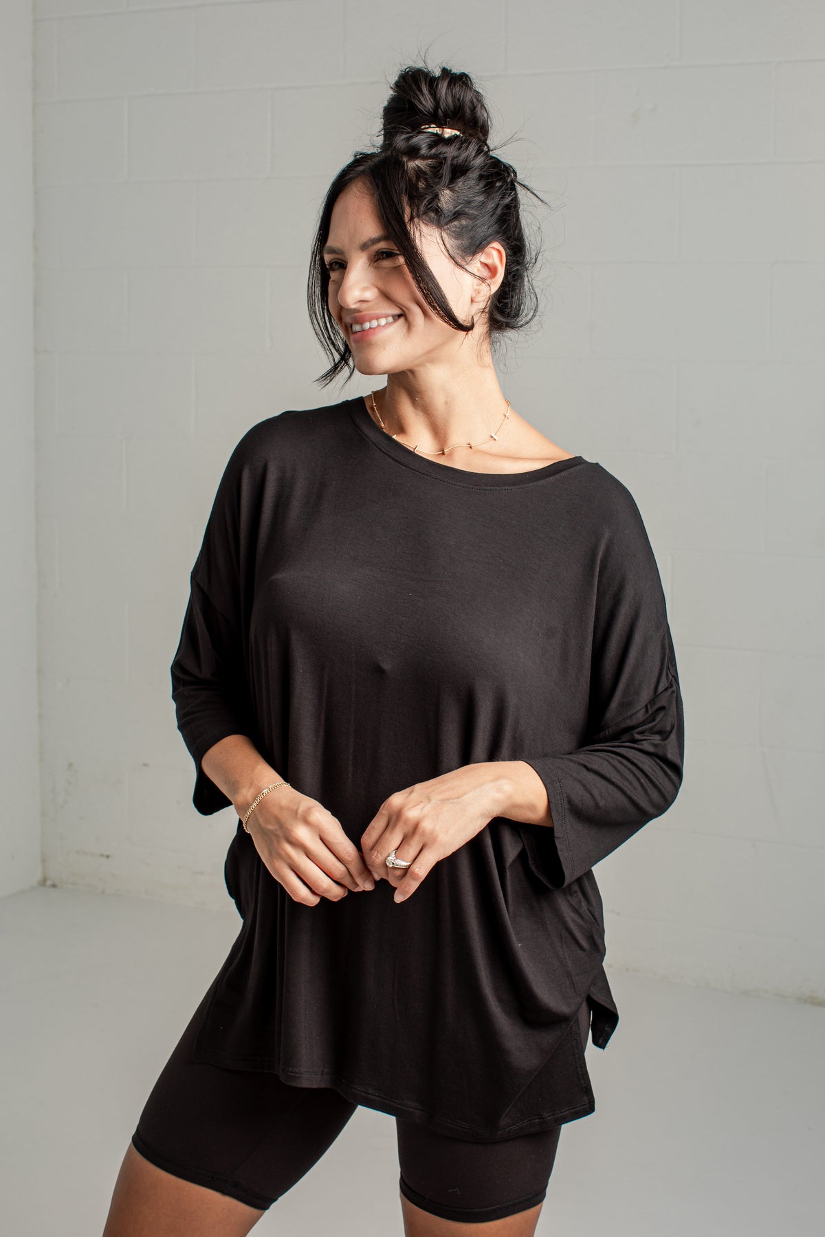 A woman wearing a loose black long-sleeve top and black fitted shorts, standing against a light gray wall in a studio setting.