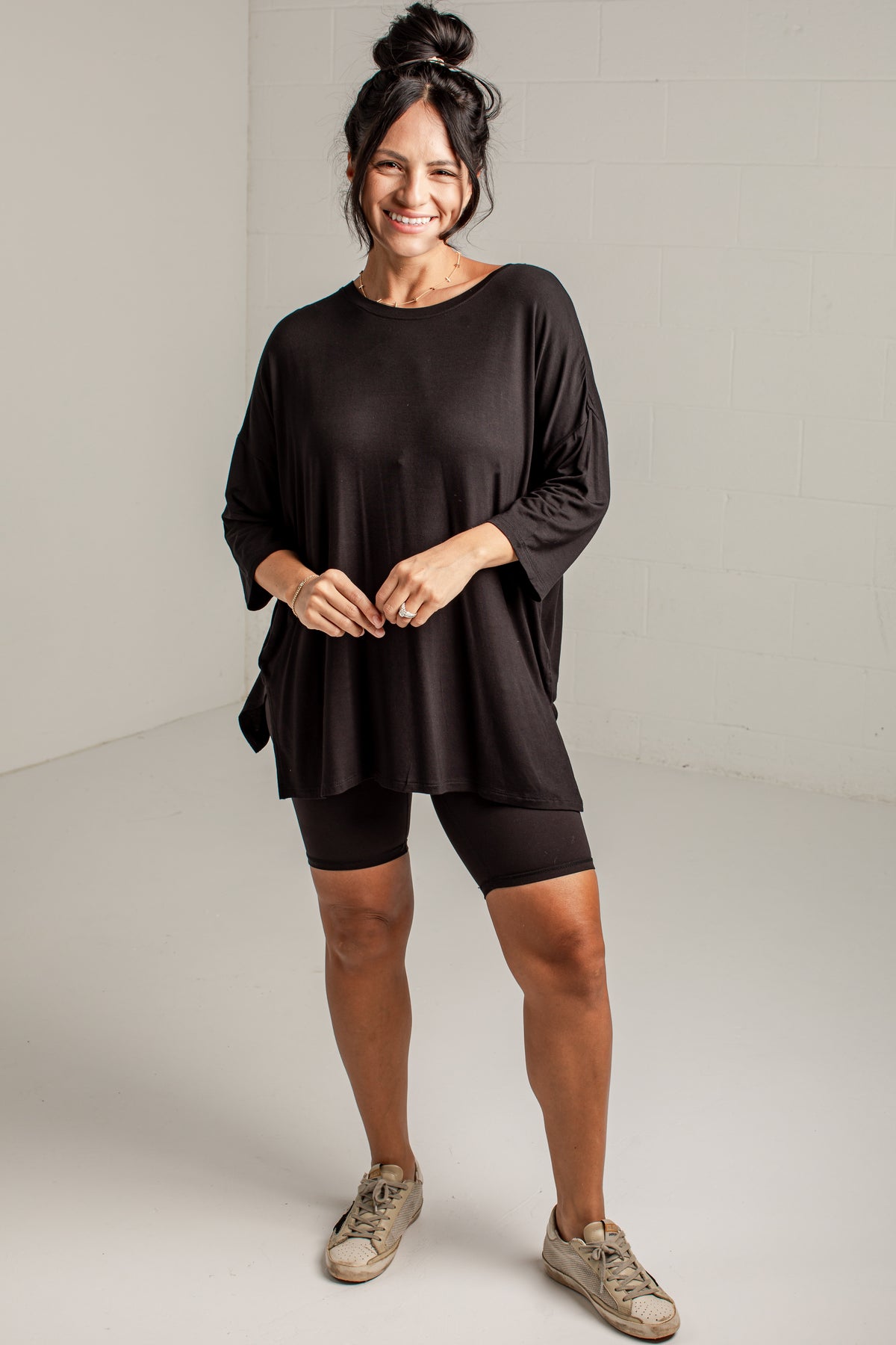 A woman modeling a black oversized three-quarter sleeve top with matching black biker shorts and beige sneakers, standing against a light gray studio wall.